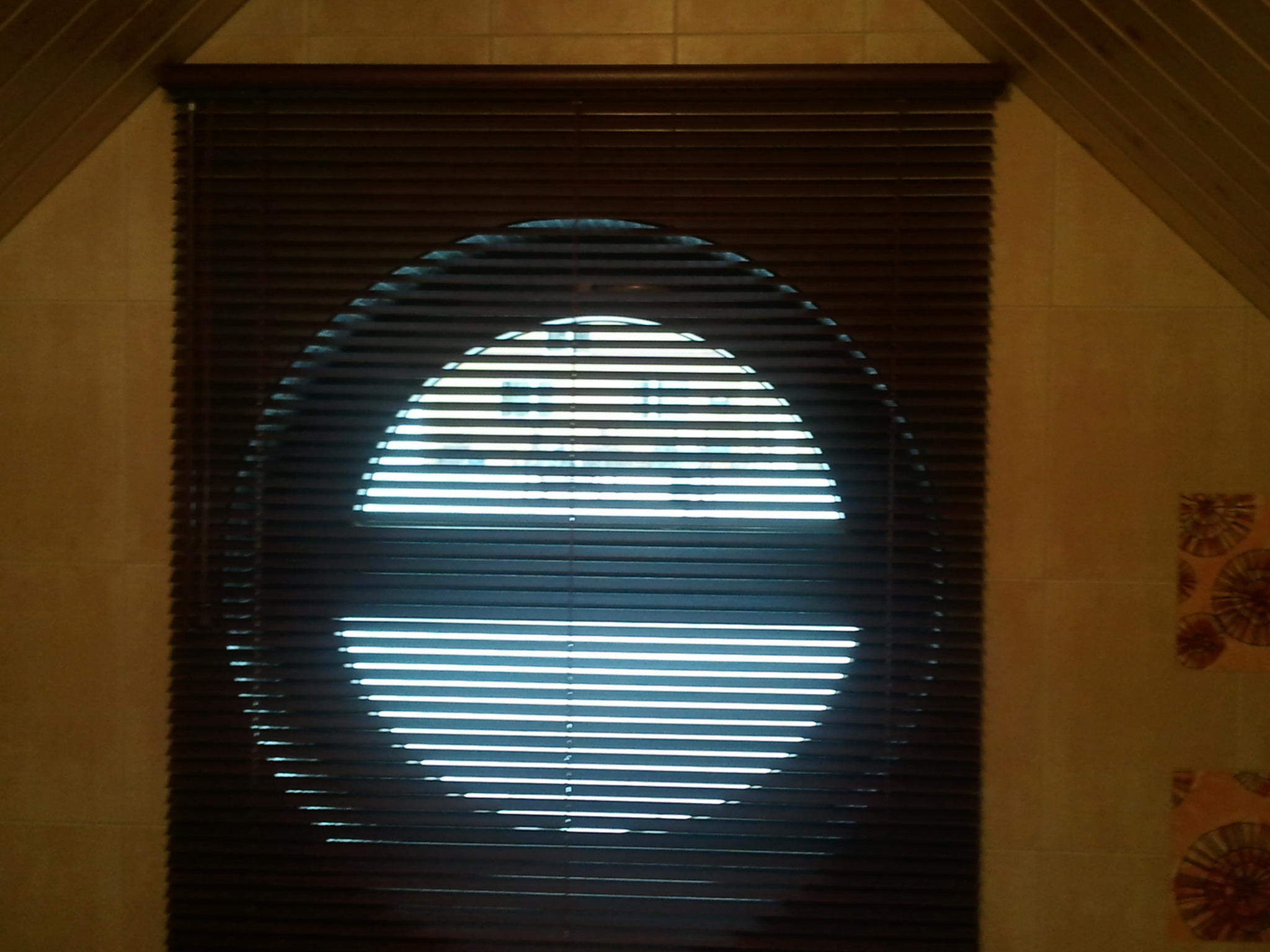 Ciemnobrązowe żaluzje aluminiowe zamontowane w oknie o nietypowym, okrągłym kształcie, wpuszczające jasne światło do wnętrza. Widoczne beżowe płytki na ścianie.