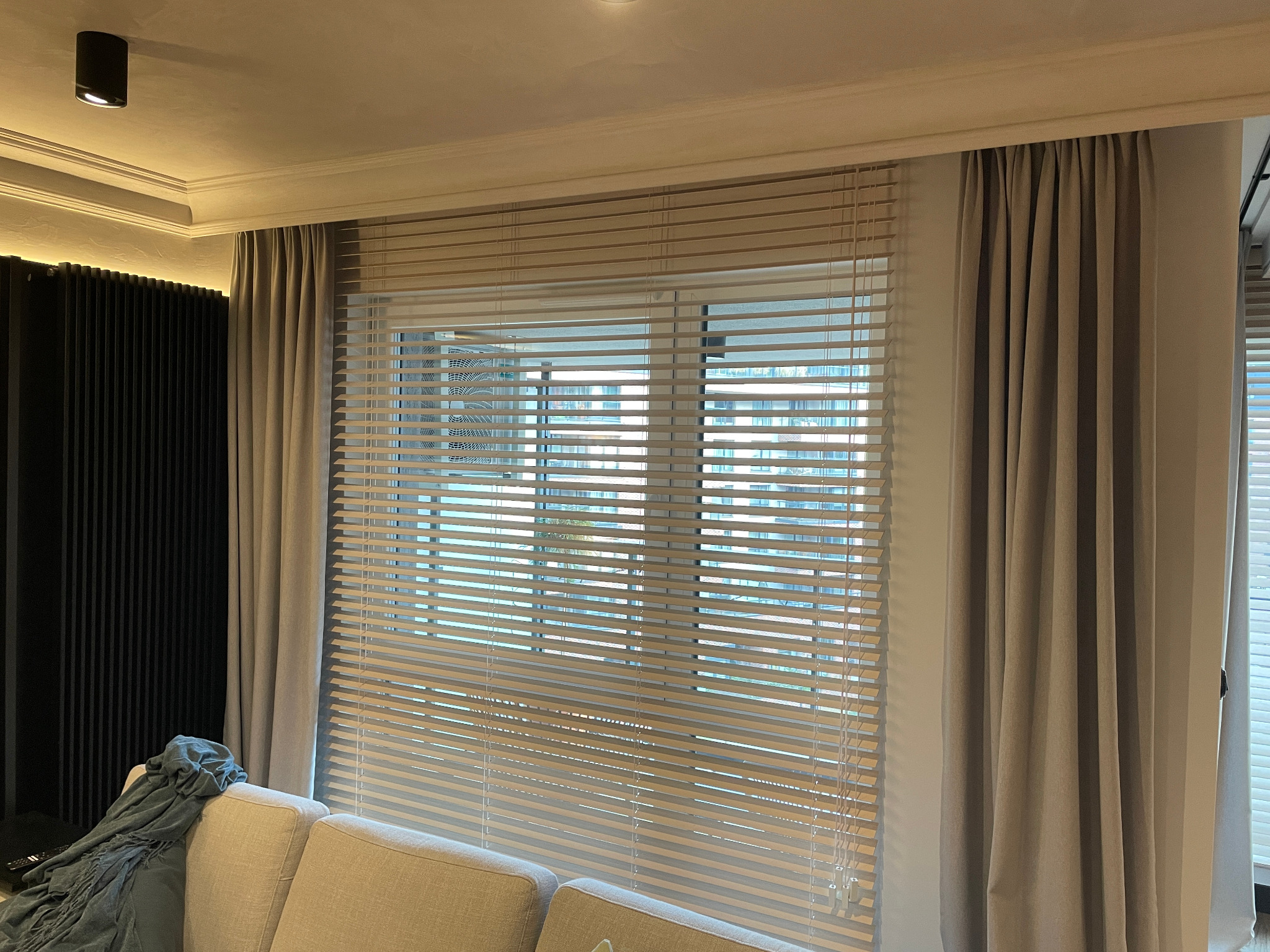 Beżowe żaluzje aluminiowe poziome zamontowane w oknie, zasłonięte częściowo beżowymi zasłonami. Widok z wnętrza pomieszczenia, w tle zabudowa ściany.