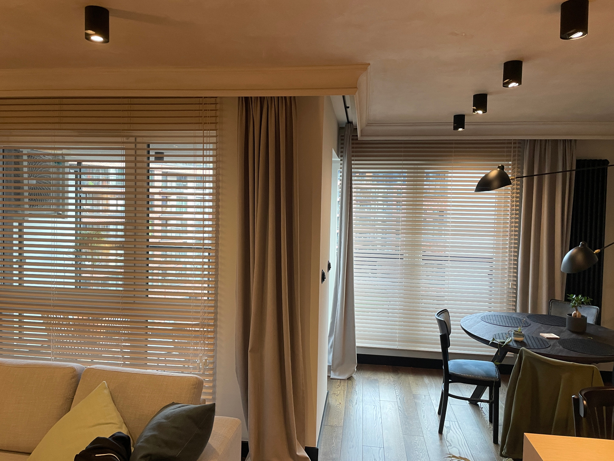 Jasne, drewniane żaluzje poziome w nowoczesnym salonie z beżową sofą i zasłonami. Widoczne oświetlenie sufitowe i stół z krzesłami.