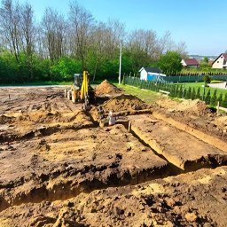 Fenomen usługi ziemne - Żółta koparko-ładowarka przy wykopie pod fundamenty domu jednorodzinnego. Widok z góry na wykopane rowy w ziemi, w tle zieleń i budynek.