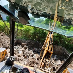 Fenomen usługi ziemne - Widok z kabiny koparki na wykop pełen gruzu i ziemi, z zielenią w tle. Operator w kabinie, widoczne elementy sterowania i fragment nogi. Nietypowa perspektywa.