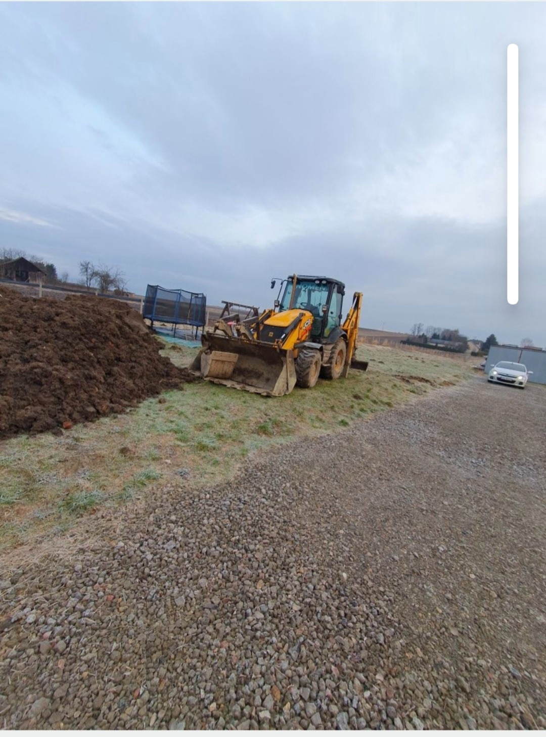 Żółta koparko-ładowarka JCB na polu z pryzmą ziemi i utwardzoną drogą. W tle dom, trampolina i auto. Pochmurne niebo.