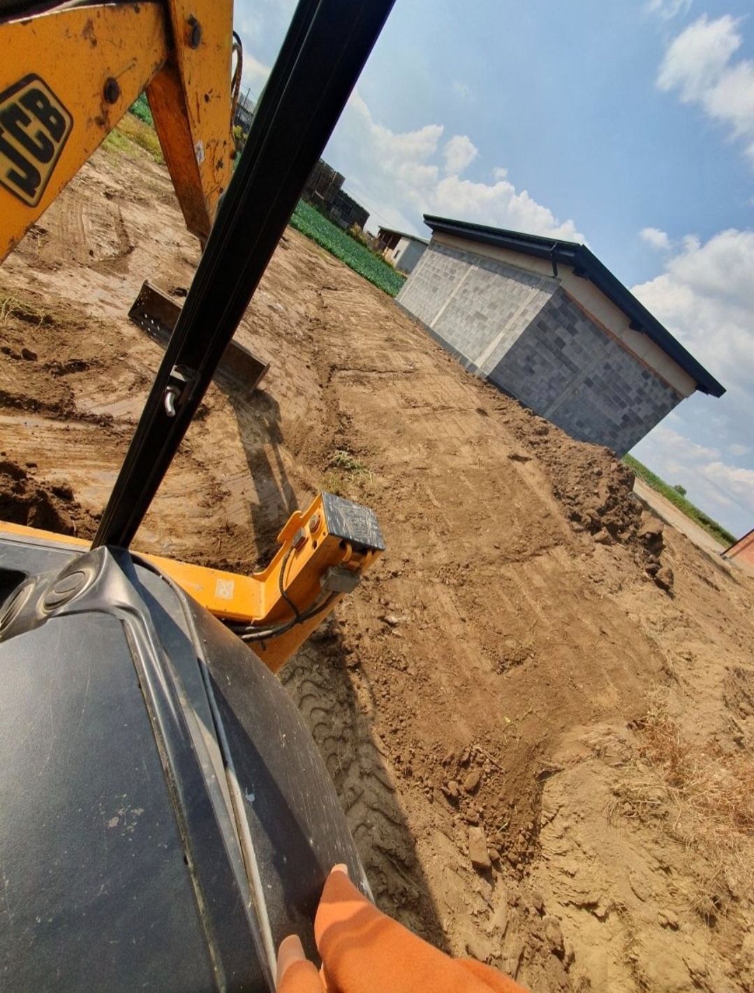 Żółta koparka JCB na nierównym, brązowym terenie budowy z budynkiem w tle, widziana z perspektywy operatora. Ślady opon na ziemi. Błękitne niebo z chmurami.