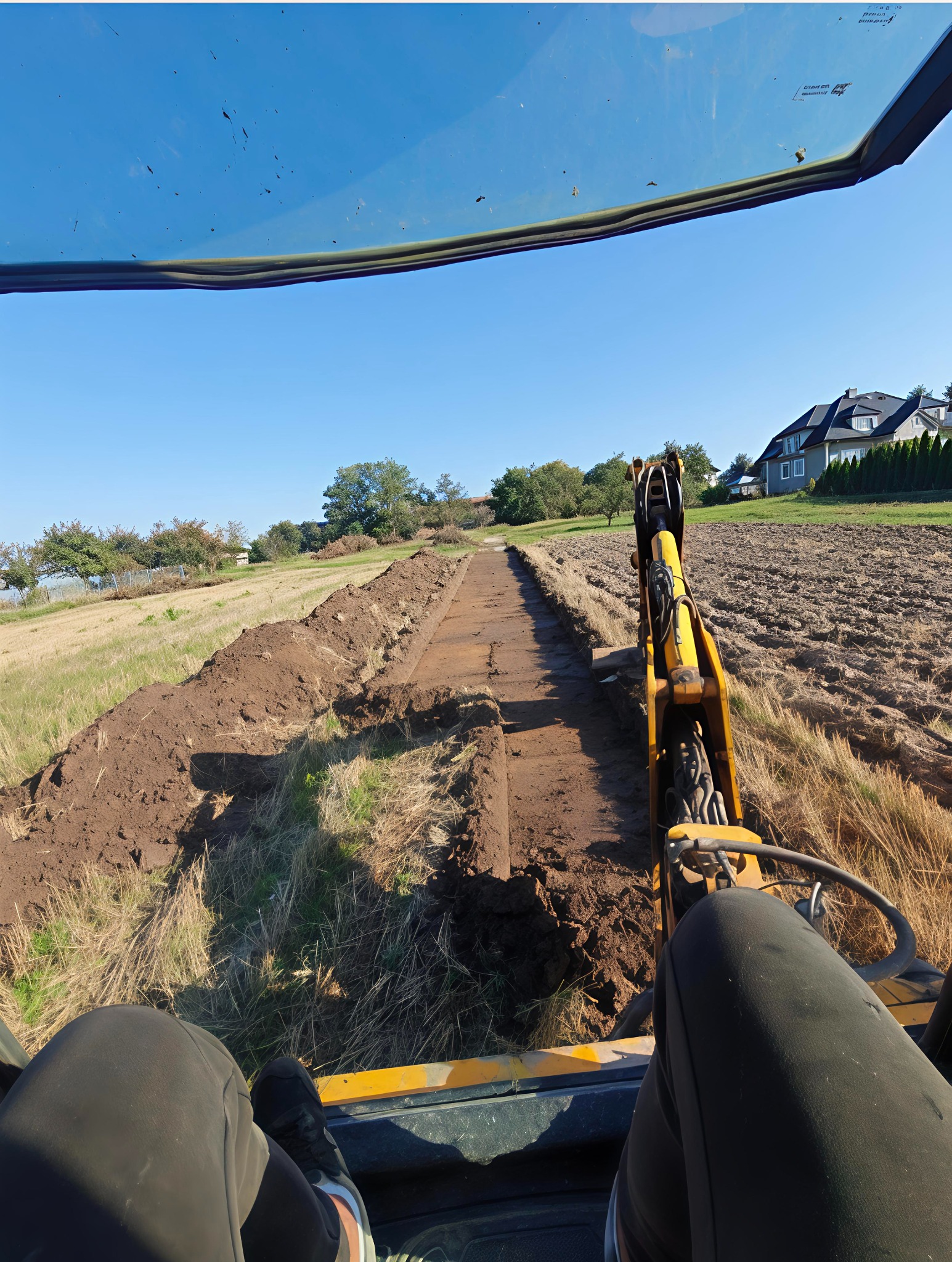 Wykop pod instalacje widziany z kabiny koparki, równy rów na polu, w tle dom i drzewa, słoneczny dzień.