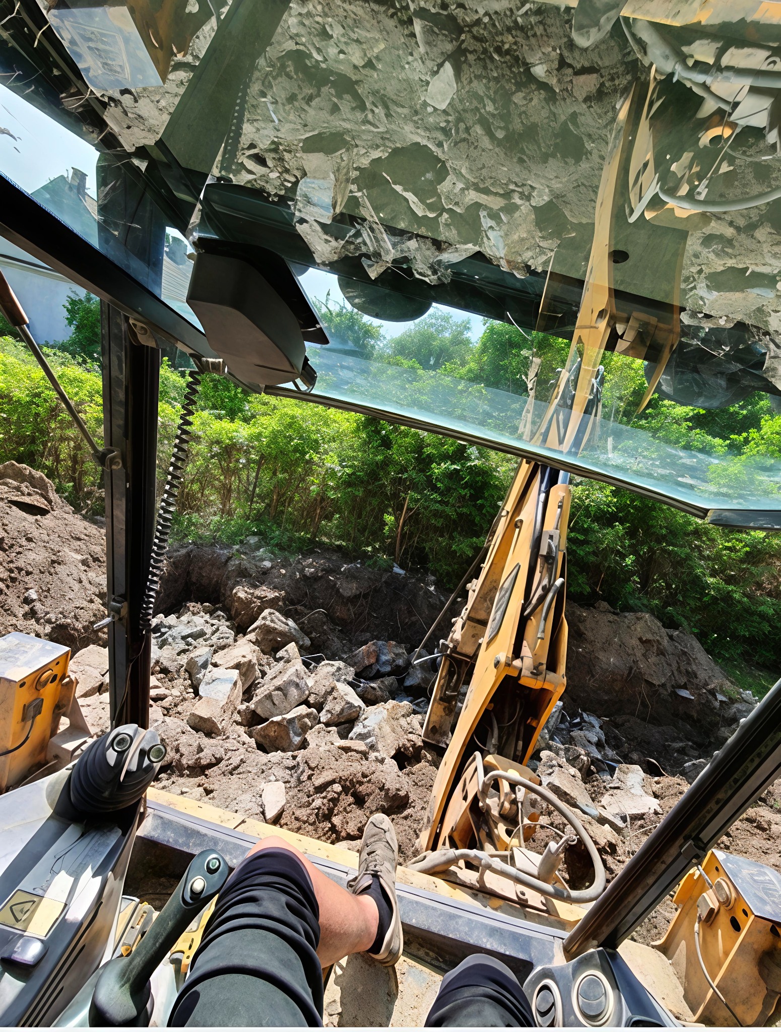 Widok z kabiny koparki na wykop pełen gruzu i ziemi, z zielenią w tle. Operator w kabinie, widoczne elementy sterowania i fragment nogi. Nietypowa perspektywa.