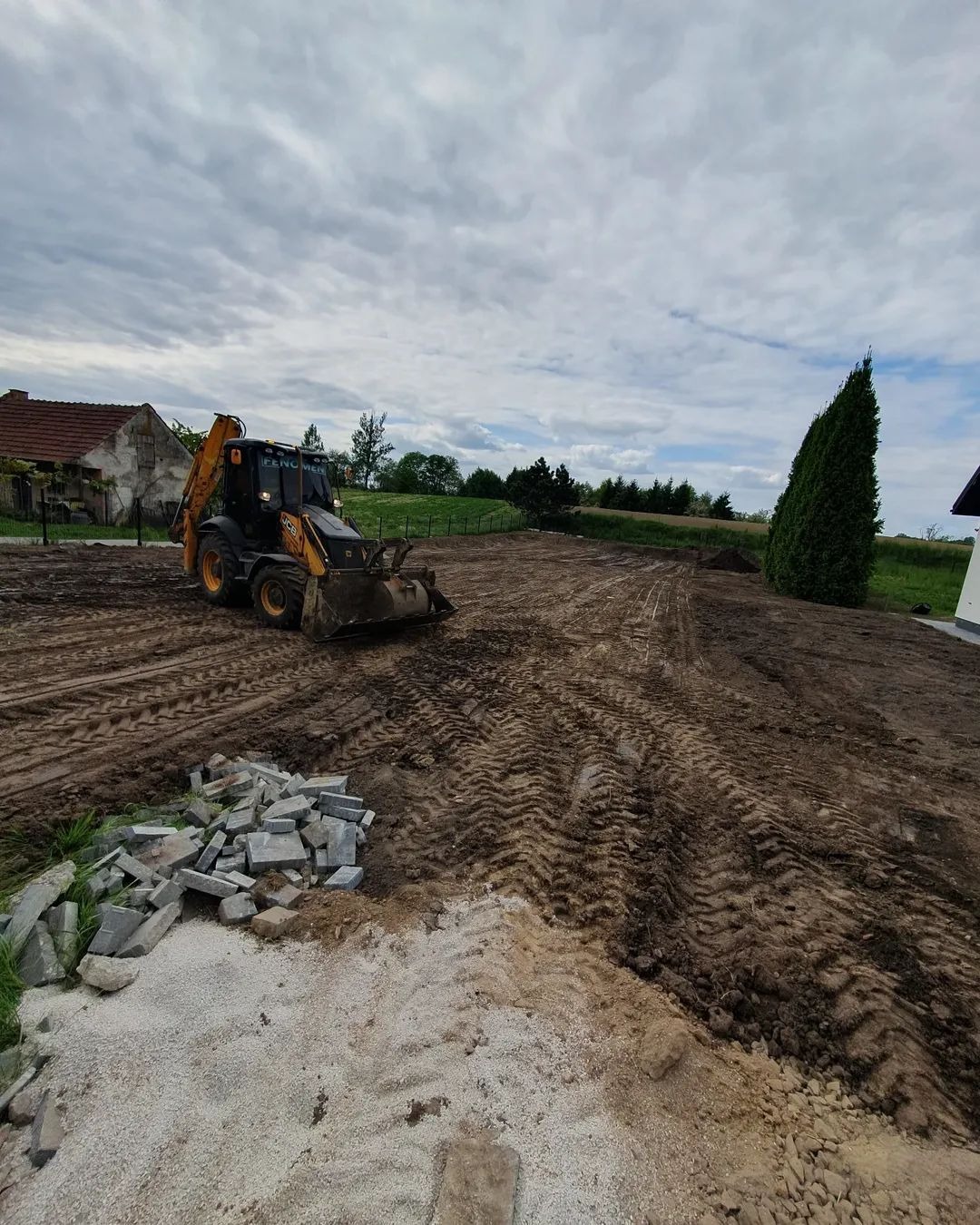 Żółto-czarna koparko-ładowarka na wyrównanym, brązowym terenie z widocznymi śladami opon. W tle dom z czerwoną dachówką i zielone drzewa pod pochmurnym niebem.