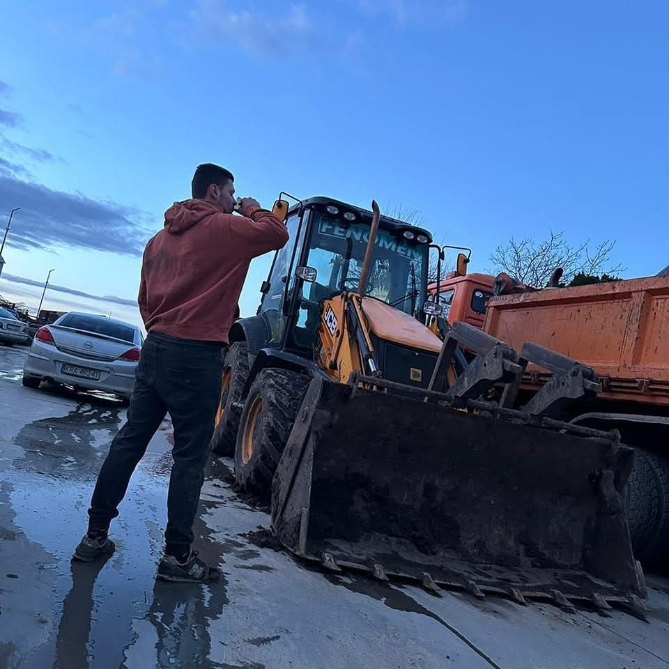 Operator pije przed koparko-ładowarką JCB z brudną łyżką, obok wywrotka. W tle zaparkowane auto i pochmurne niebo. Przerwa w pracy na budowie.