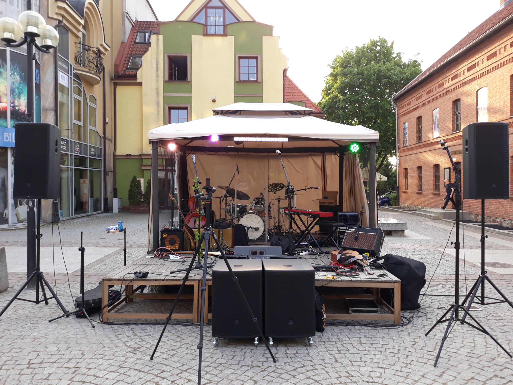 Scena plenerowa z nagłośnieniem i instrumentami muzycznymi pod namiotem, przygotowana do występu na brukowanym placu miejskim.
