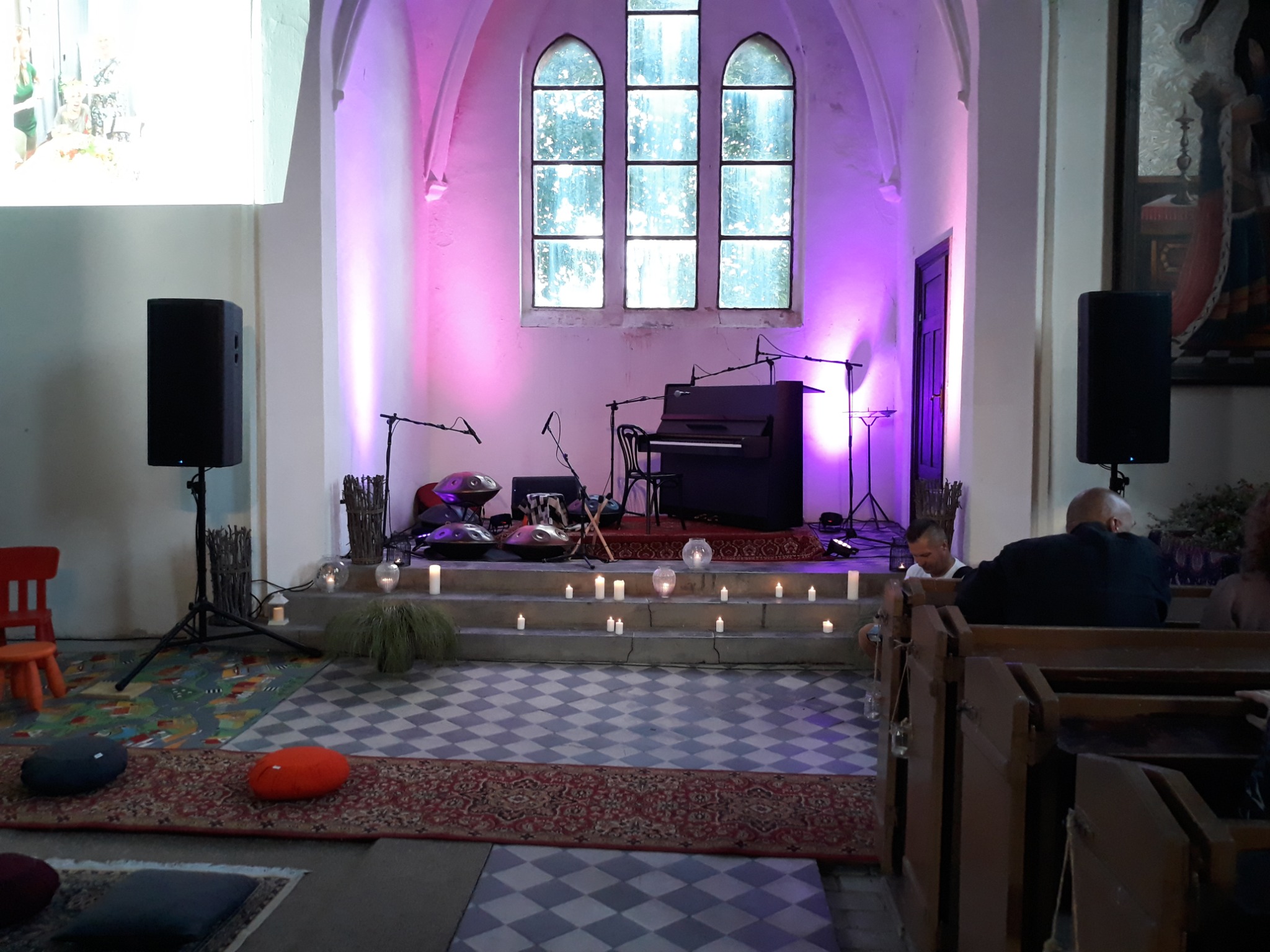 Kościół zaadaptowany na salę koncertową: scena z pianinem, handpanami, świecami i nagłośnieniem, oświetlona fioletowym światłem. Widzowie siedzą w ławkach.