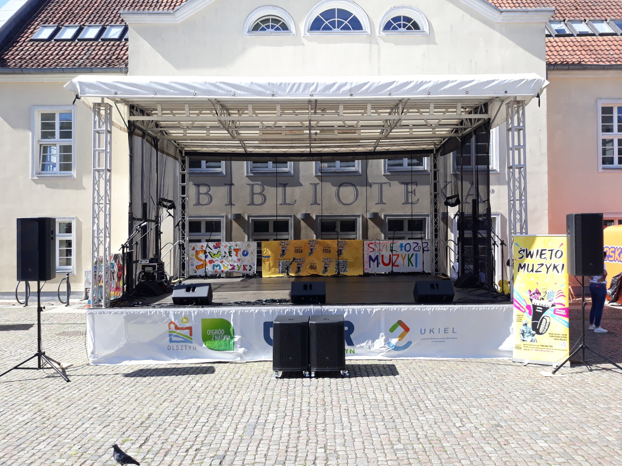 Scena plenerowa z nagłośnieniem i banerami 'Święto Muzyki' na tle budynku Biblioteki w Olsztynie. Widoczne głośniki, mikrofony i elementy konstrukcji scenicznej.