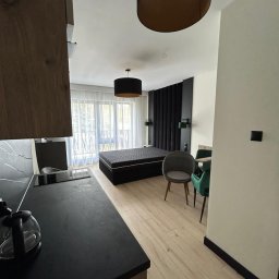 Well Done - Nowoczesne wnętrze apartamentu: czarna zabudowa kuchenna z blatem imitującym kamień, drewniana podłoga i sypialnia z czarnym zagłówkiem. Widok z perspektywy kuchni.