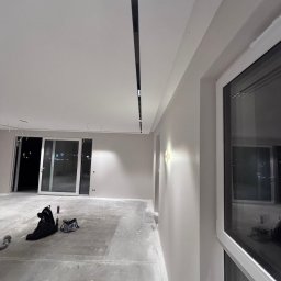 Well Done - Wnętrze w trakcie prac wykończeniowych: świeżo pomalowane ściany w odcieniach szarości, sufit z zamontowanym oświetleniem liniowym, duże okno z widokiem na miasto.