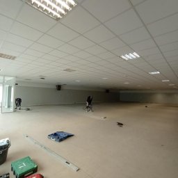 Well Done - Duża przestrzeń biurowa w trakcie prac wykończeniowych: szlifowanie posadzki przez dwóch pracowników, widoczne narzędzia Hilti i poziomica. Oświetlenie sufitowe.