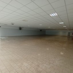 Well Done - Duża, pusta hala biurowa z beżową podłogą z płytek, szarymi ścianami i białym sufitem kasetonowym. Widoczne klimatyzatory i przeszklone drzwi wejściowe.