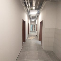 Well Done - Długi korytarz po remoncie z szarymi płytkami, białymi ścianami i brązowymi drzwiami. Widoczne instalacje pod sufitem i fragment ściany z płytek. Oświetlenie sufitowe.