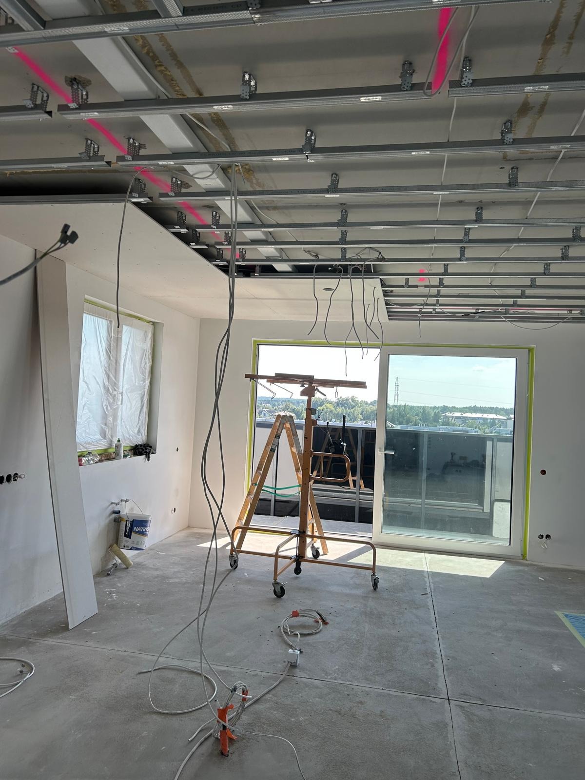 Montaż sufitu podwieszanego z płyt karton-gips: widoczne profile, płyty w trakcie instalacji, podnośnik i przewody elektryczne. Jasne wnętrze z oknem balkonowym.