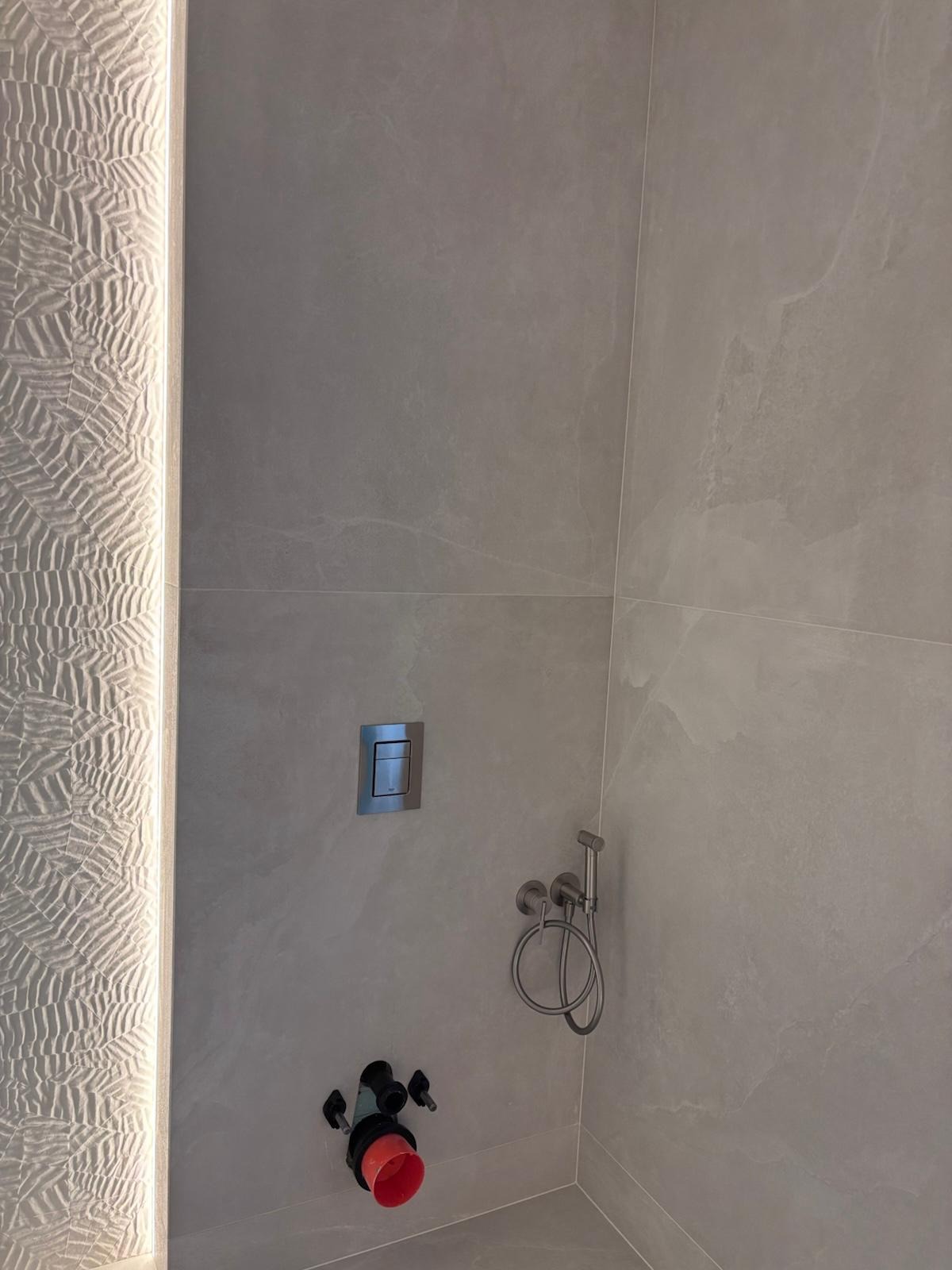 Wykończona łazienka z szarymi płytkami imitującymi kamień, ozdobnym oświetleniem LED i zamontowaną armaturą. Widoczne przyłącza do toalety i bidetki.