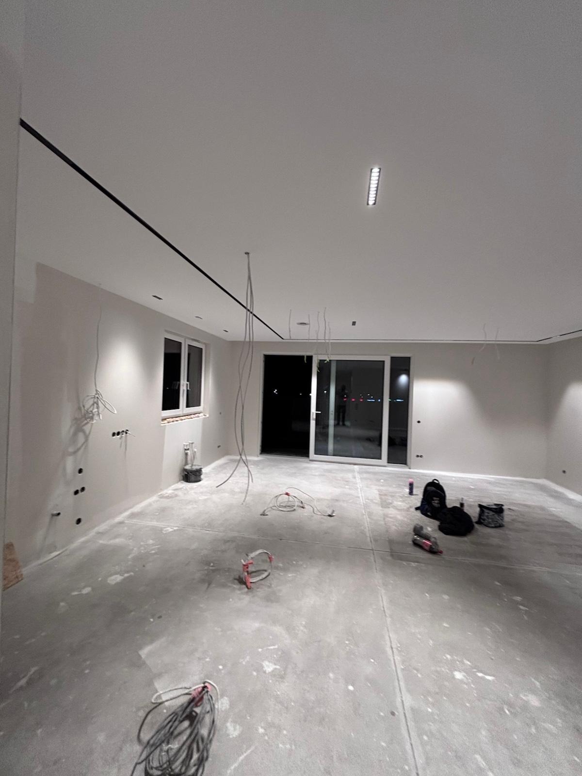 Wnętrze w trakcie prac wykończeniowych: widoczne przewody elektryczne, gładkie ściany, surowa podłoga, okno i przesuwne drzwi balkonowe. Minimalistyczny design.