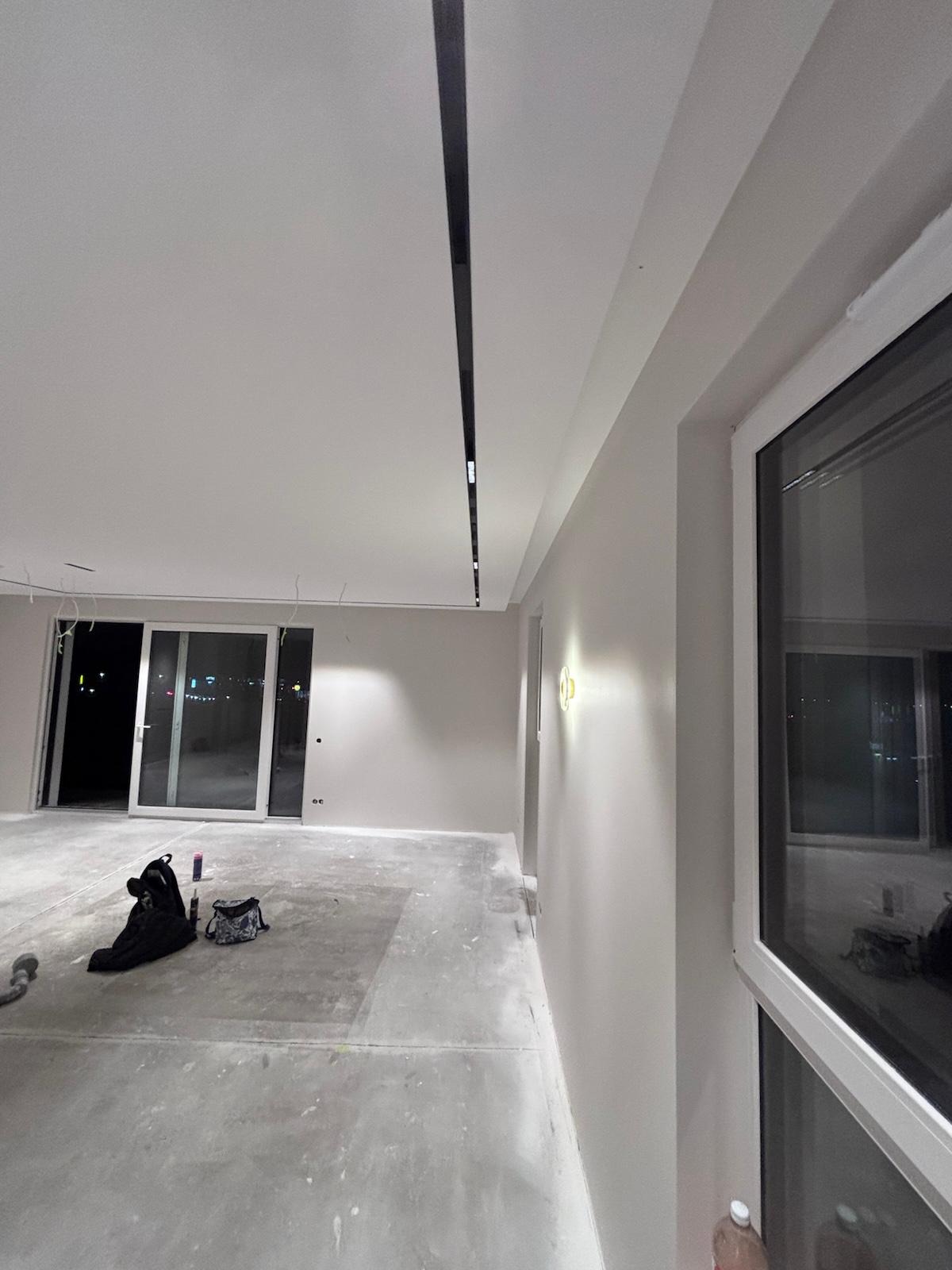 Wnętrze w trakcie prac wykończeniowych: świeżo pomalowane ściany w odcieniach szarości, sufit z zamontowanym oświetleniem liniowym, duże okno z widokiem na miasto.