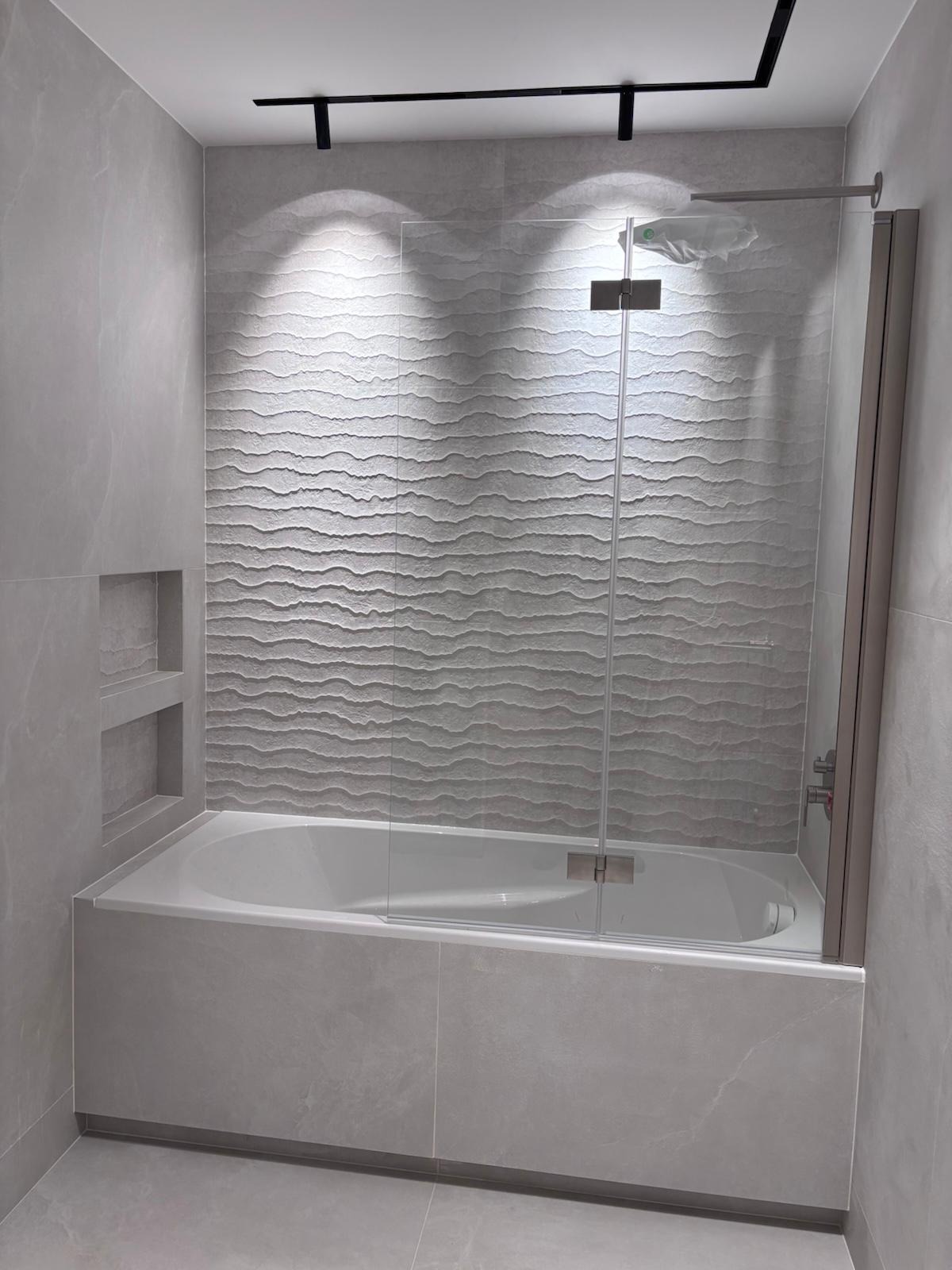 Wykończona łazienka w odcieniach szarości z wanną obudowaną płytkami, szklanym parawanem nawannowym i dekoracyjną, falistą ścianą z płytek. Nowoczesne oświetlenie sufitowe.