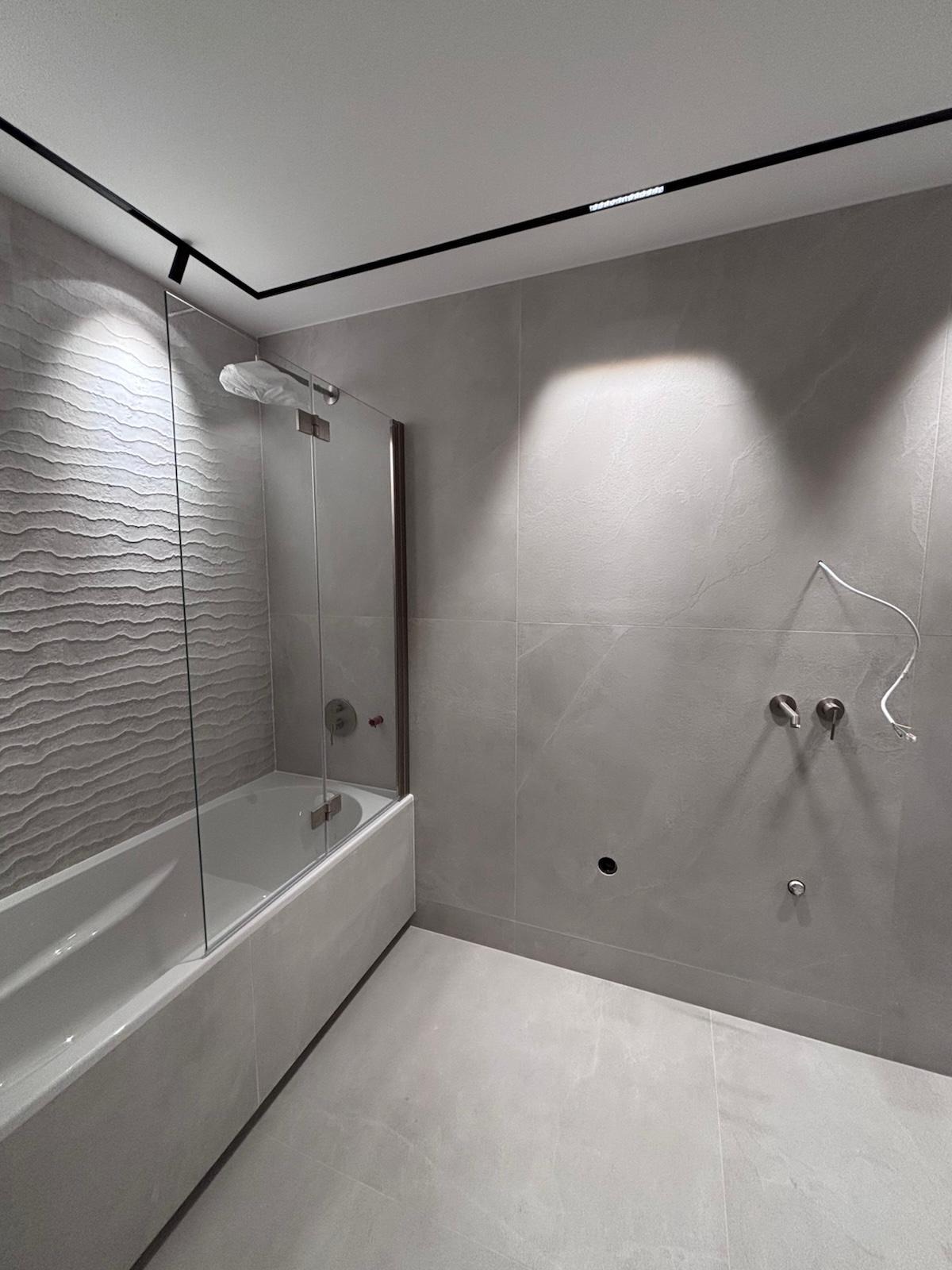 Nowoczesna łazienka w szarościach: płytki imitujące marmur, kabina nawannowa ze szkła, minimalistyczne oświetlenie sufitowe i armatura podtynkowa.