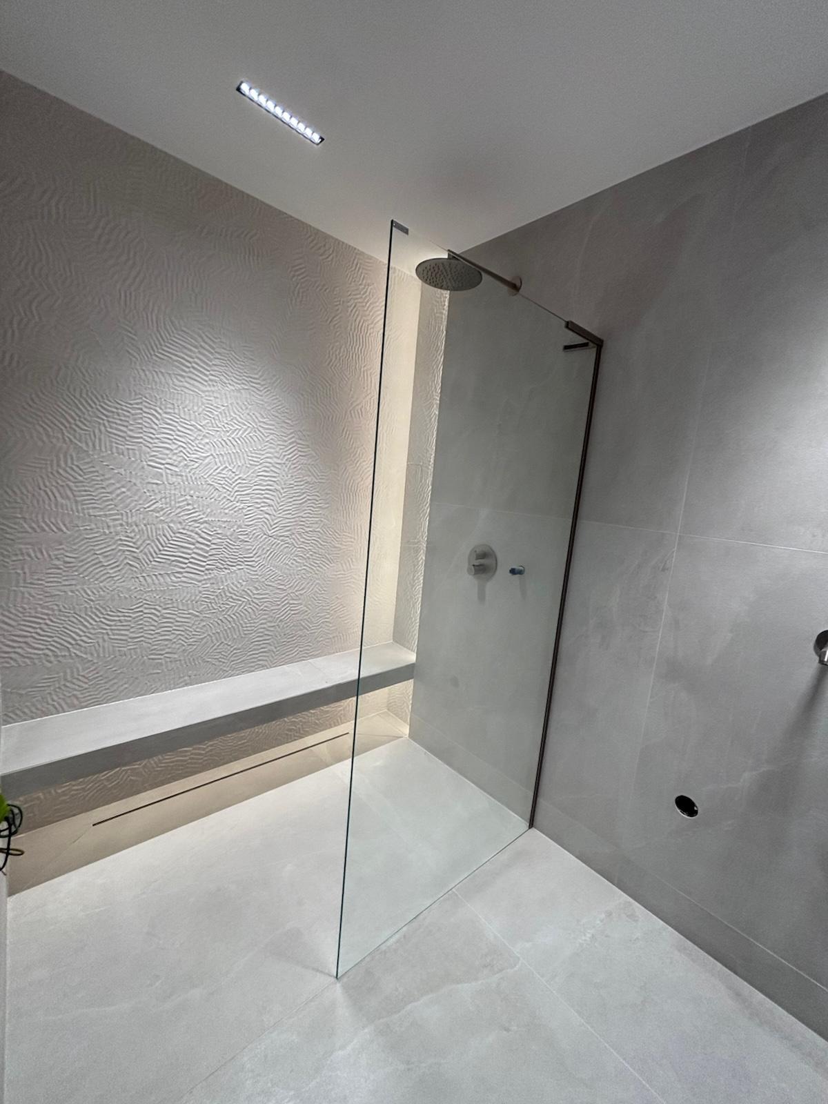 Nowoczesna łazienka w odcieniach szarości z kabiną prysznicową typu walk-in, podświetlaną półką i dekoracyjną strukturą ściany. Minimalistyczny design, eleganckie wykończenie.