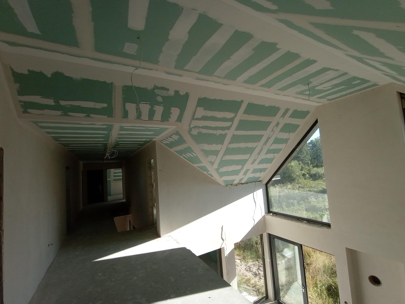 Sufit z płyt kartonowo-gipsowych w surowym wnętrzu z dużym oknem. Widoczne łączenia płyt i fragmenty instalacji elektrycznej. W tle korytarz w stanie deweloperskim.