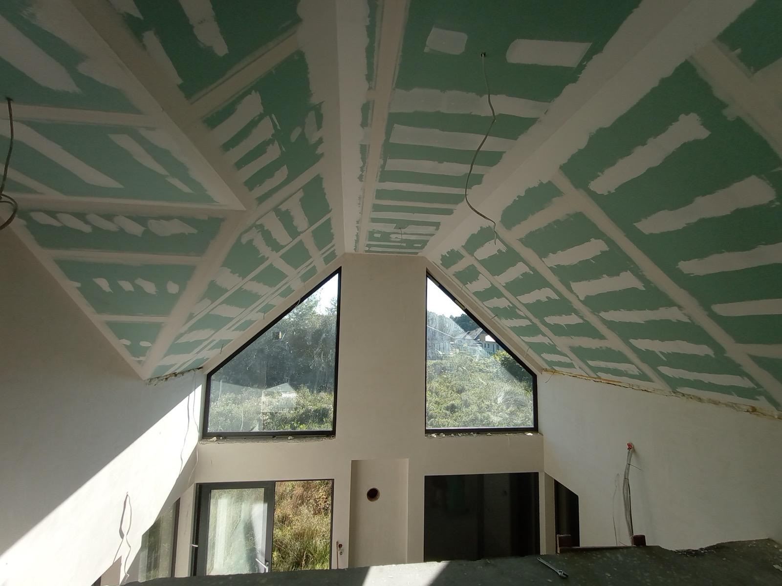Skośny sufit poddasza z zamontowaną płytą karton-gips, widoczne łączenia i szpachlowanie, okna trójkątne w szczycie, surowe wykończenie wnętrza.