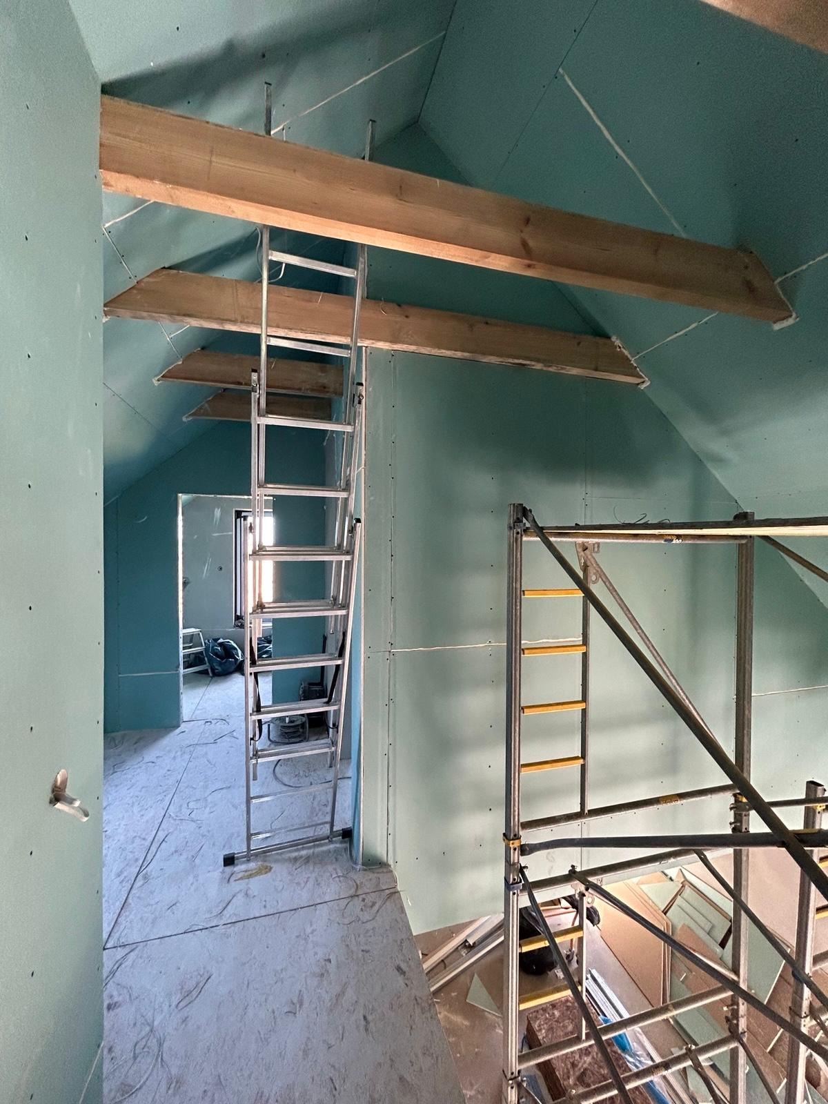 Poddasze z płytami gipsowo-kartonowymi w trakcie montażu, widoczne drewniane belki, drabina i rusztowanie. Ściany w kolorze jasnej zieleni.