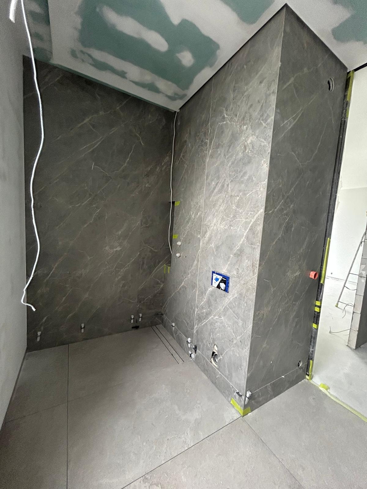 Wykończona łazienka z szarymi płytkami imitującymi marmur na ścianach i podłodze. Widoczne przygotowane instalacje wodne i elektryczne. Sufit z płyt kartonowo-gipsowych.
