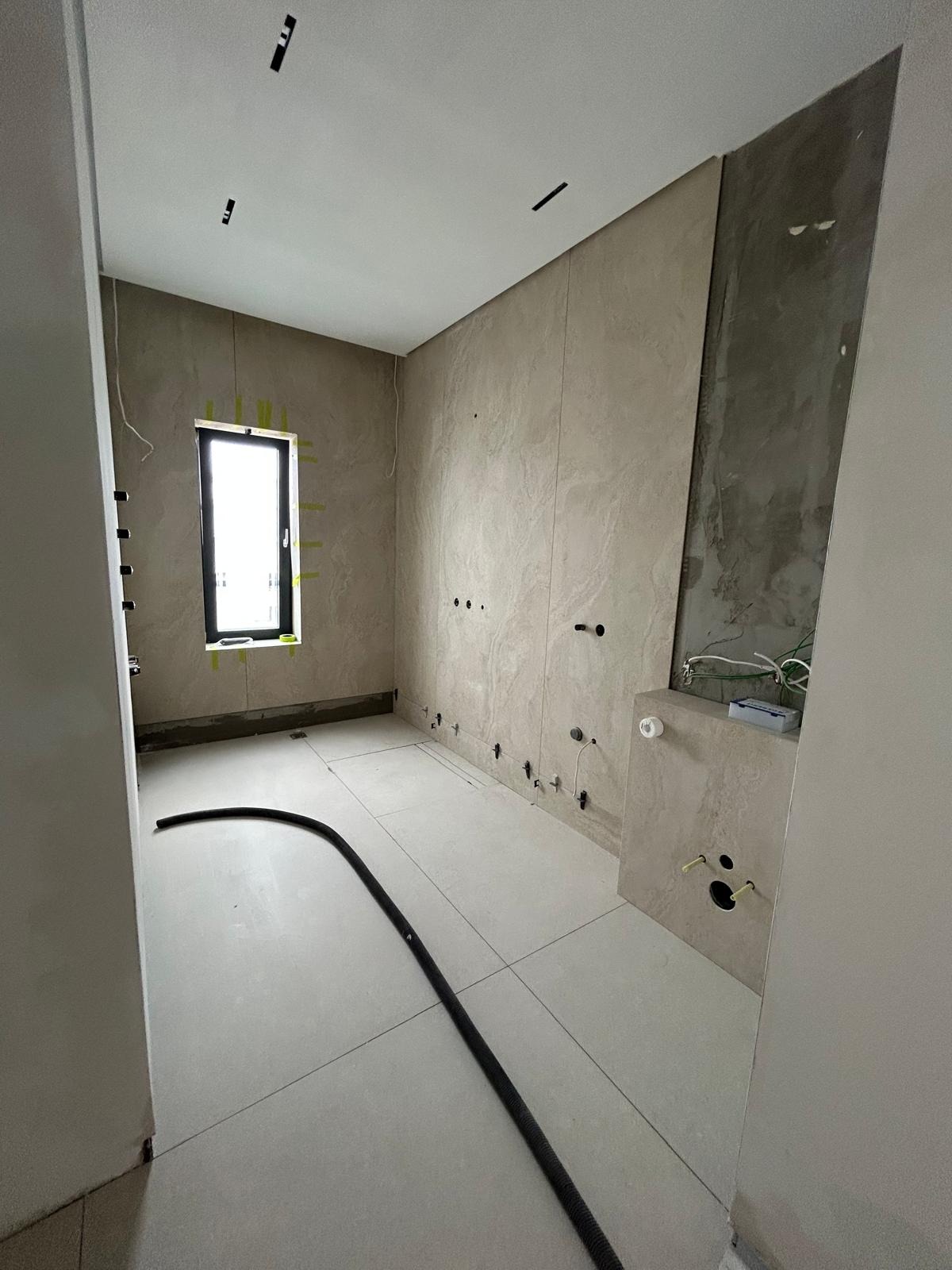 Wnętrze łazienki w trakcie wykończenia: beżowe płytki na ścianach i podłodze, widoczne instalacje, okno z czarną ramą. Częściowo położone płytki, surowy stan.