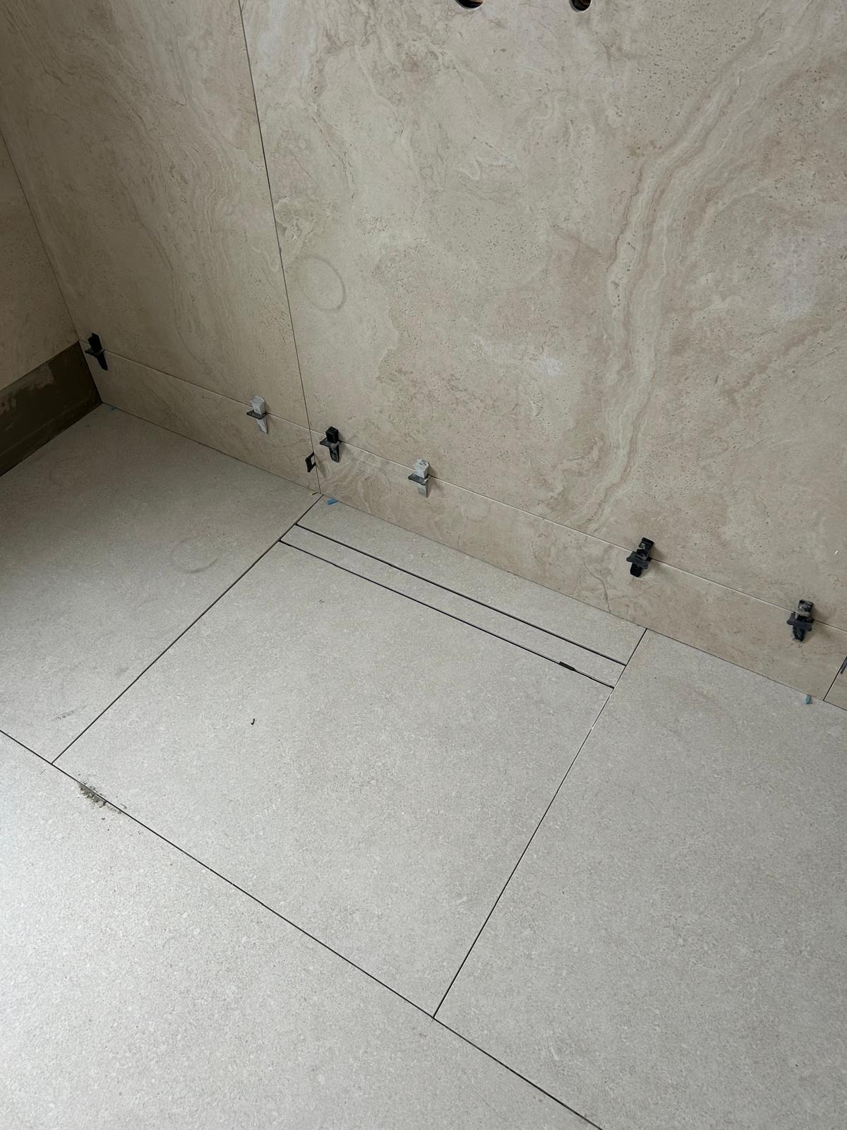 Układanie beżowych płytek na podłodze i ścianie z użyciem klipsów poziomujących. Widoczne fugi i fragmenty kleju. Praca w toku, etap wykończeniowy.