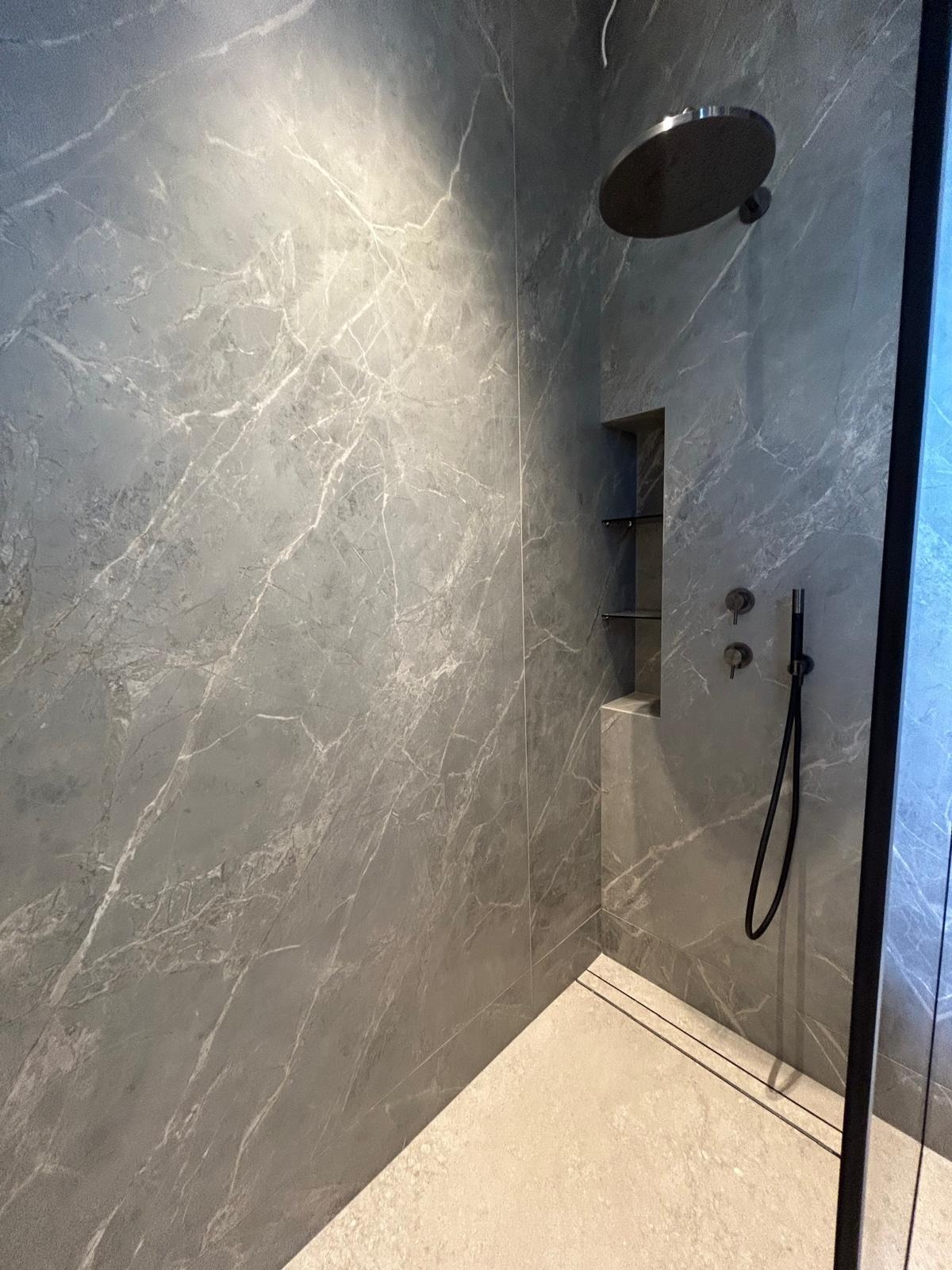 Nowoczesna kabina prysznicowa z szarymi płytkami imitującymi marmur, deszczownicą i wnęką z półkami. Minimalistyczny design, eleganckie wykończenie.