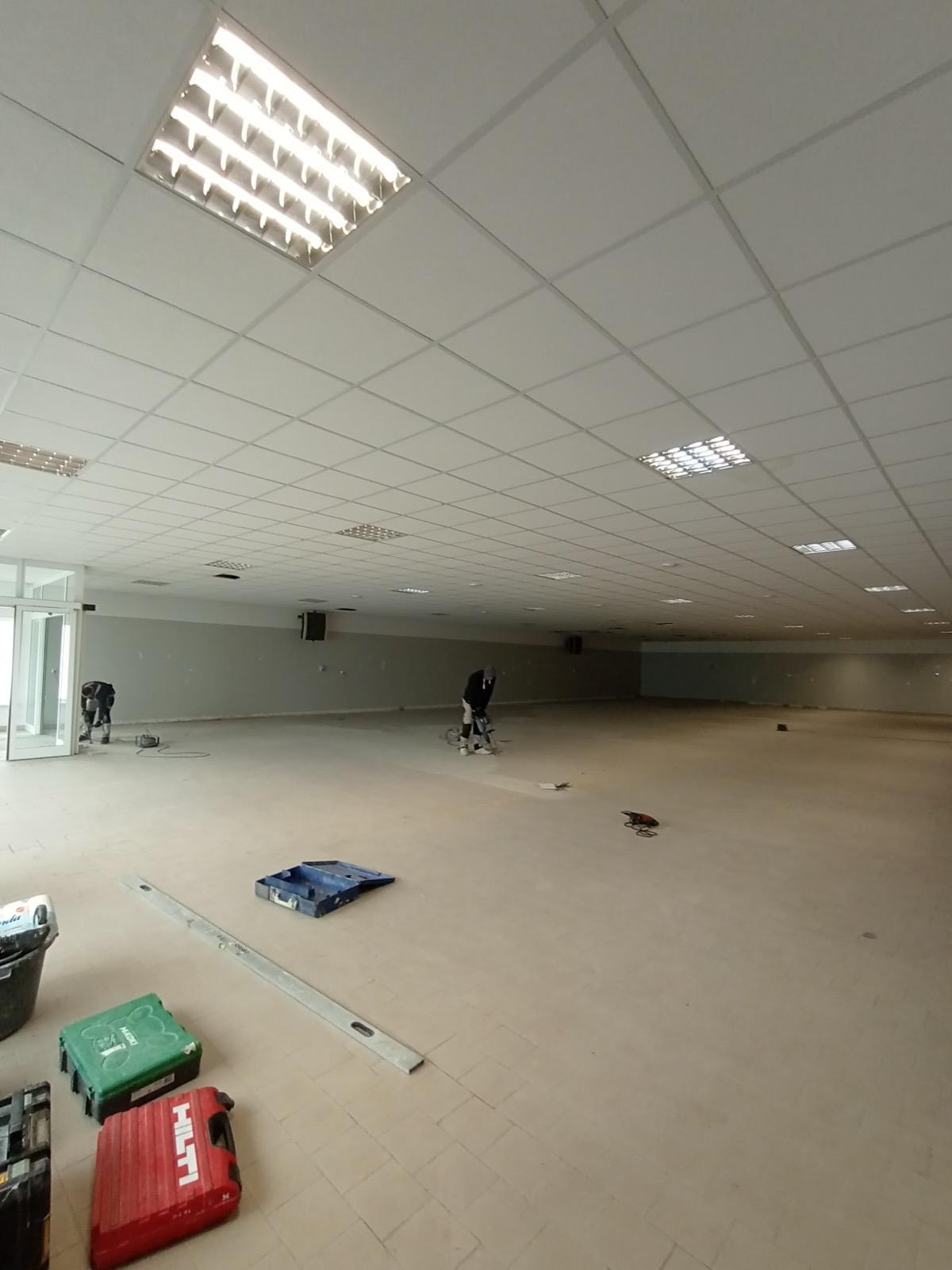 Duża przestrzeń biurowa w trakcie prac wykończeniowych: szlifowanie posadzki przez dwóch pracowników, widoczne narzędzia Hilti i poziomica. Oświetlenie sufitowe.
