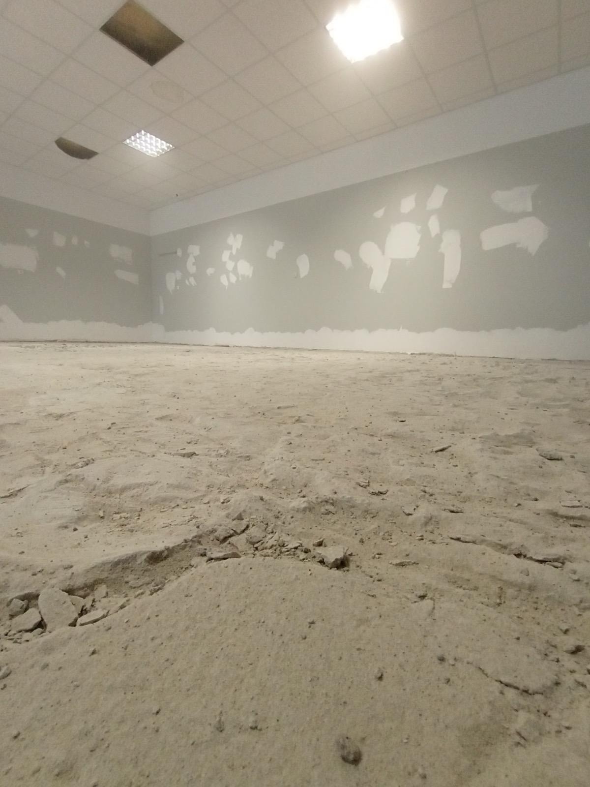 Wnętrze w trakcie prac przygotowawczych: ściany z fragmentami gładzi, surowa podłoga. Widoczne otwory w suficie pod instalacje. Ujęcie z niskiej perspektywy.