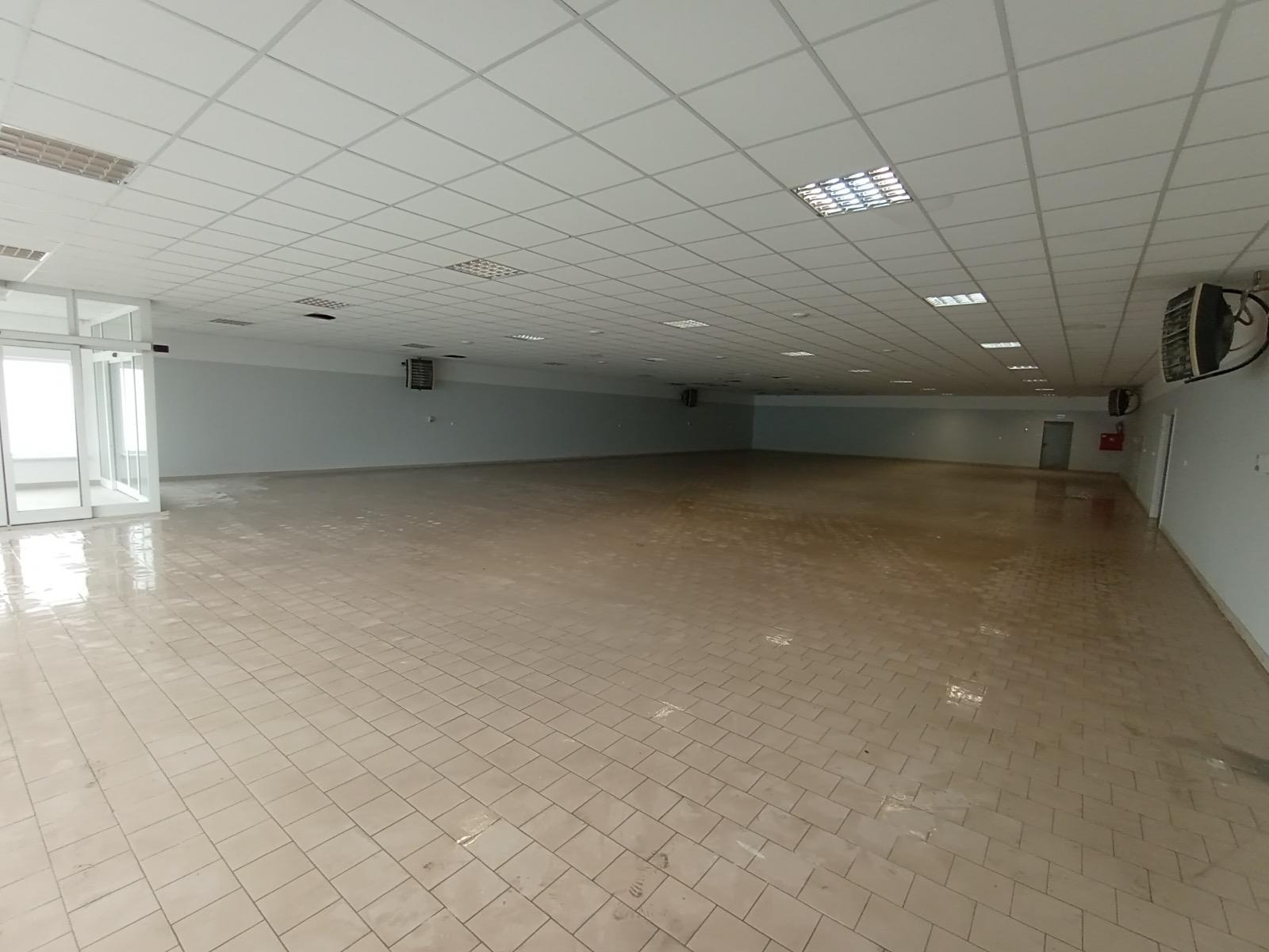 Duża, pusta hala biurowa z beżową podłogą z płytek, szarymi ścianami i białym sufitem kasetonowym. Widoczne klimatyzatory i przeszklone drzwi wejściowe.
