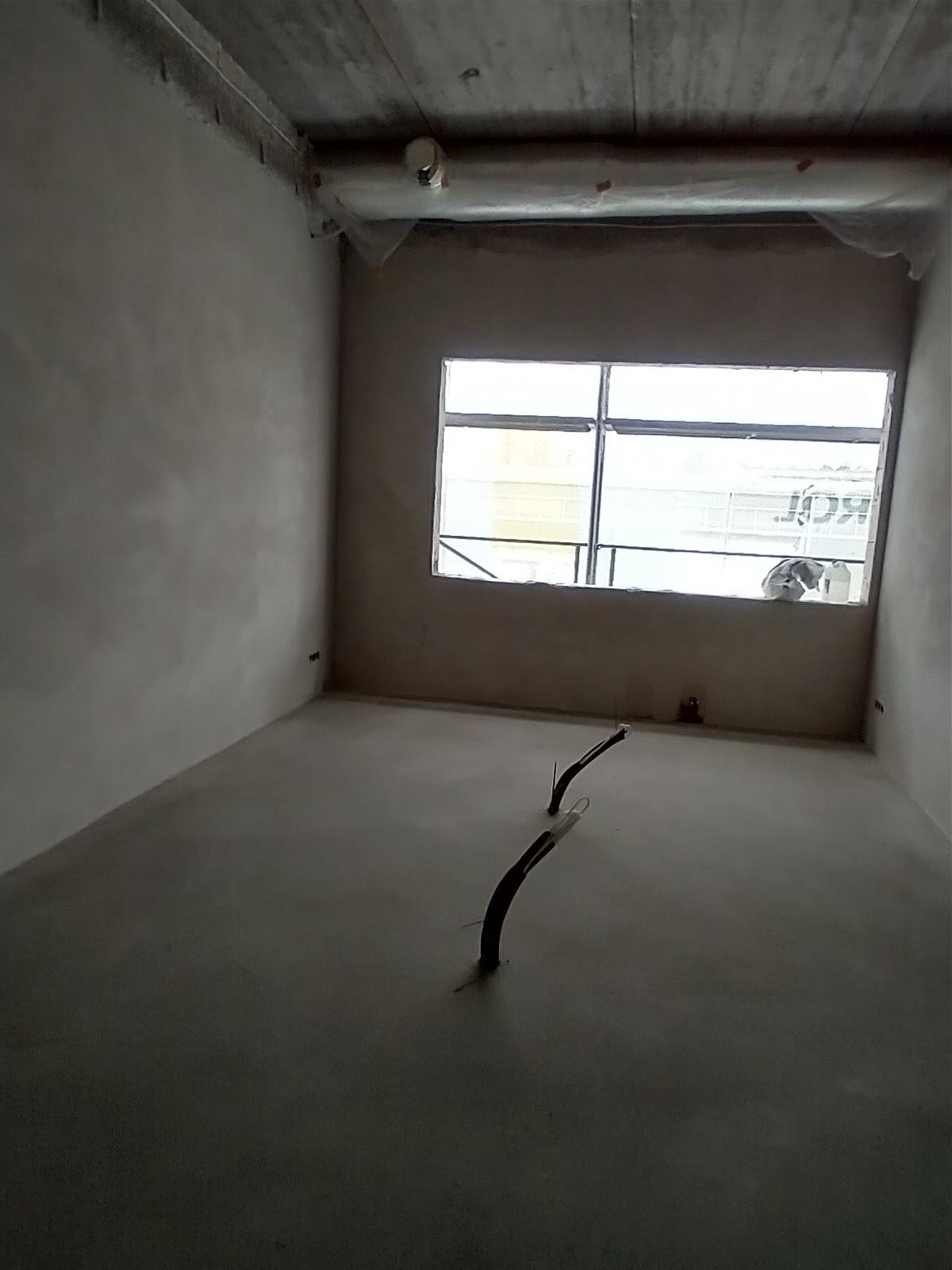 Surowe wnętrze po szpachlowaniu ścian i wylewce, widoczne wystające kable z podłogi. Okno z widokiem na budynek. Betonowy sufit z instalacją.