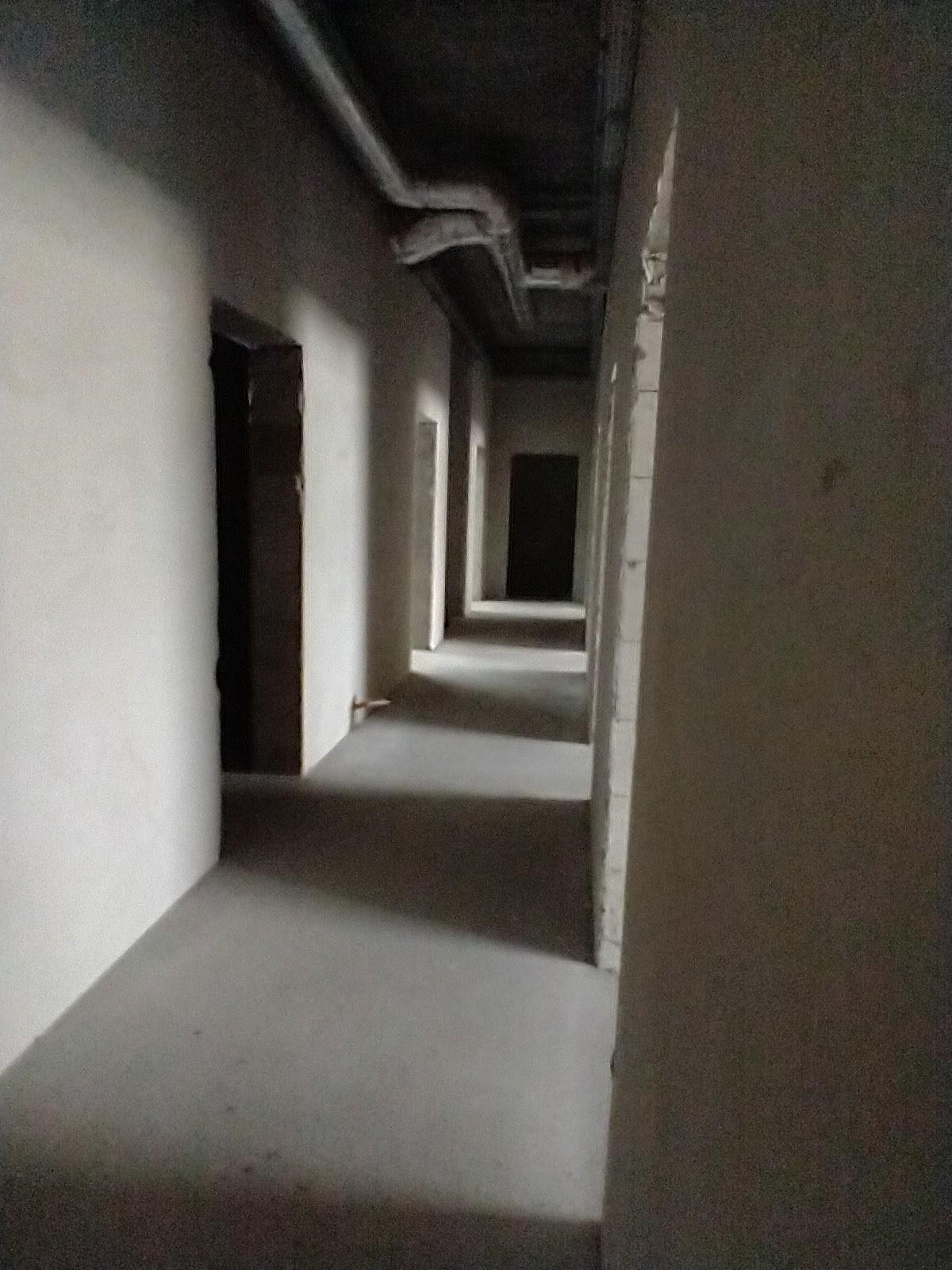 Długi korytarz w stanie surowym, ściany pokryte szpachlą, widoczne instalacje pod sufitem i otwory drzwiowe. Perspektywa z głębi korytarza.