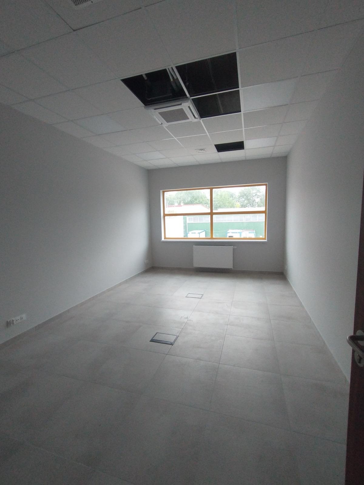Puste, jasne pomieszczenie biurowe z szarą podłogą z płytek, białymi ścianami i oknem z widokiem na zewnątrz. Sufit podwieszany z otworami.