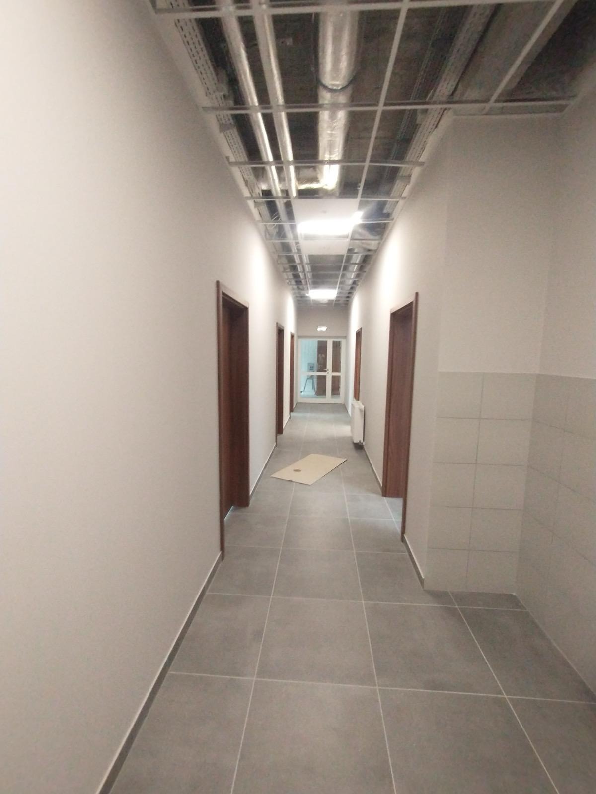 Długi korytarz po remoncie z szarymi płytkami, białymi ścianami i brązowymi drzwiami. Widoczne instalacje pod sufitem i fragment ściany z płytek. Oświetlenie sufitowe.