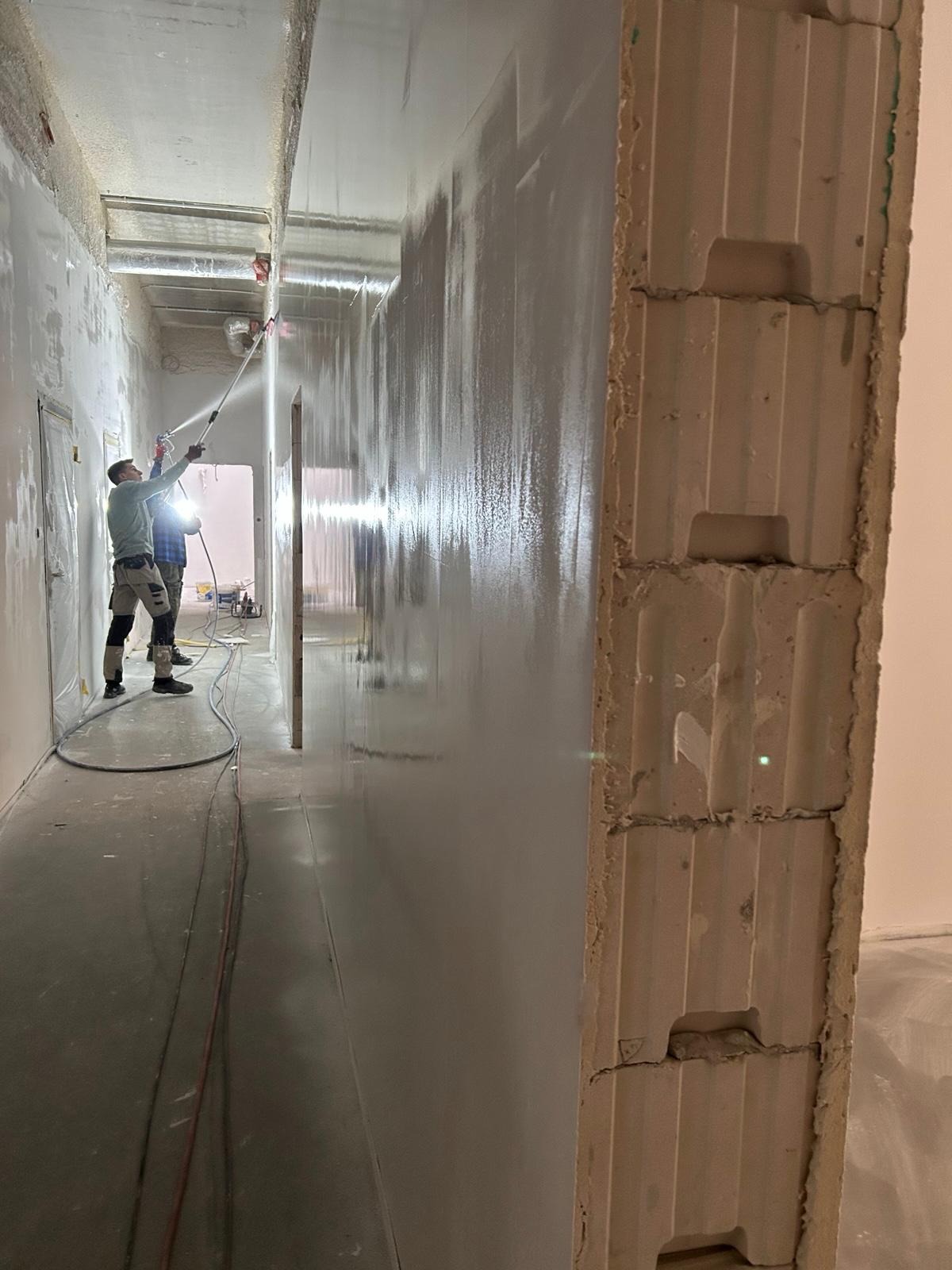 Aplikacja gładzi natryskowej w korytarzu biurowym. Dwóch pracowników w kombinezonach używa pistoletów natryskowych. Ściany pokryte świeżą, mokrą warstwą gładzi.