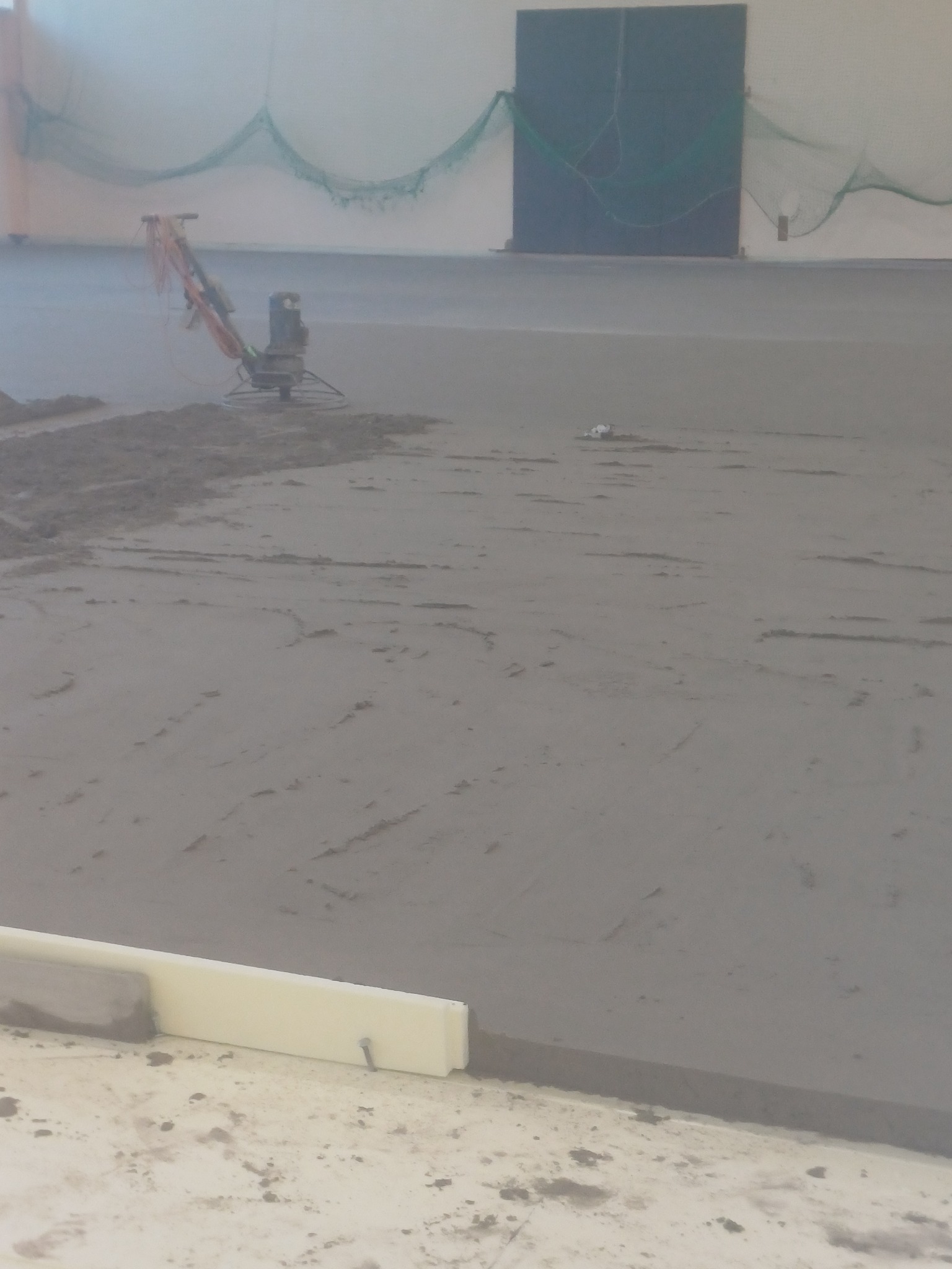 Wyrównywanie świeżej, szarej wylewki maszyną zacieraczką w hali z zieloną siatką w tle. Widoczne listwy dylatacyjne przy ścianie.