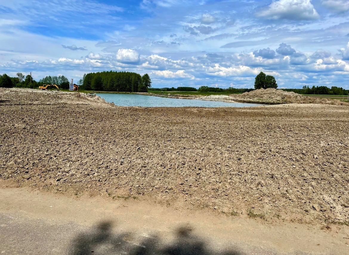 Wykopany staw w krajobrazie, z widoczną koparką na brzegu i błękitnym niebem z chmurami. Ujęcie z bliska na ziemię.