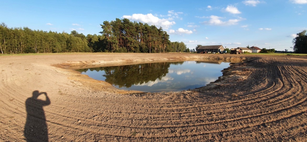 Nowo wykopany staw z wodą, otoczony świeżo wyrównanym terenem. W tle budynki gospodarcze i las. Cień fotografa w lewym dolnym rogu.