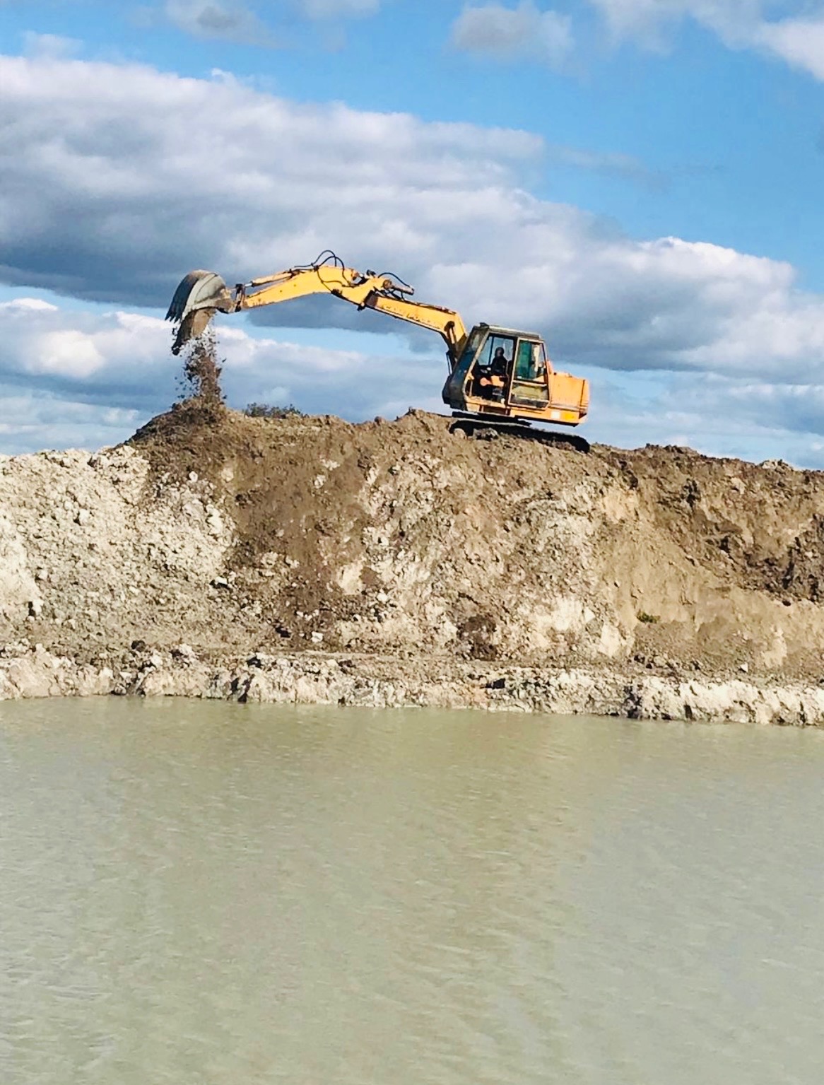 Żółta koparka nabiera ziemię z brzegu stawu na tle błękitnego nieba z chmurami. Prace ziemne, wykop.