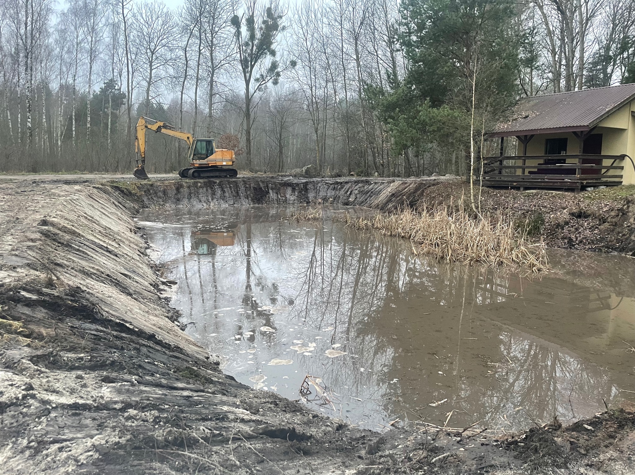 Żółta koparka przy wykopanym stawie w lesie, odbicie maszyny w wodzie. Brzegi stawu z ciemnej ziemi, w tle budynek z brązowym dachem i drzewa.