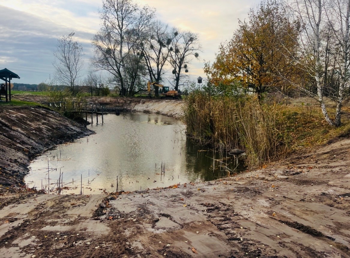 Wykończony staw z drewnianym pomostem i roślinnością brzegową. W tle widoczny sprzęt budowlany i drzewa. Ślady opon na ziemi sugerują niedawne prace ziemne.