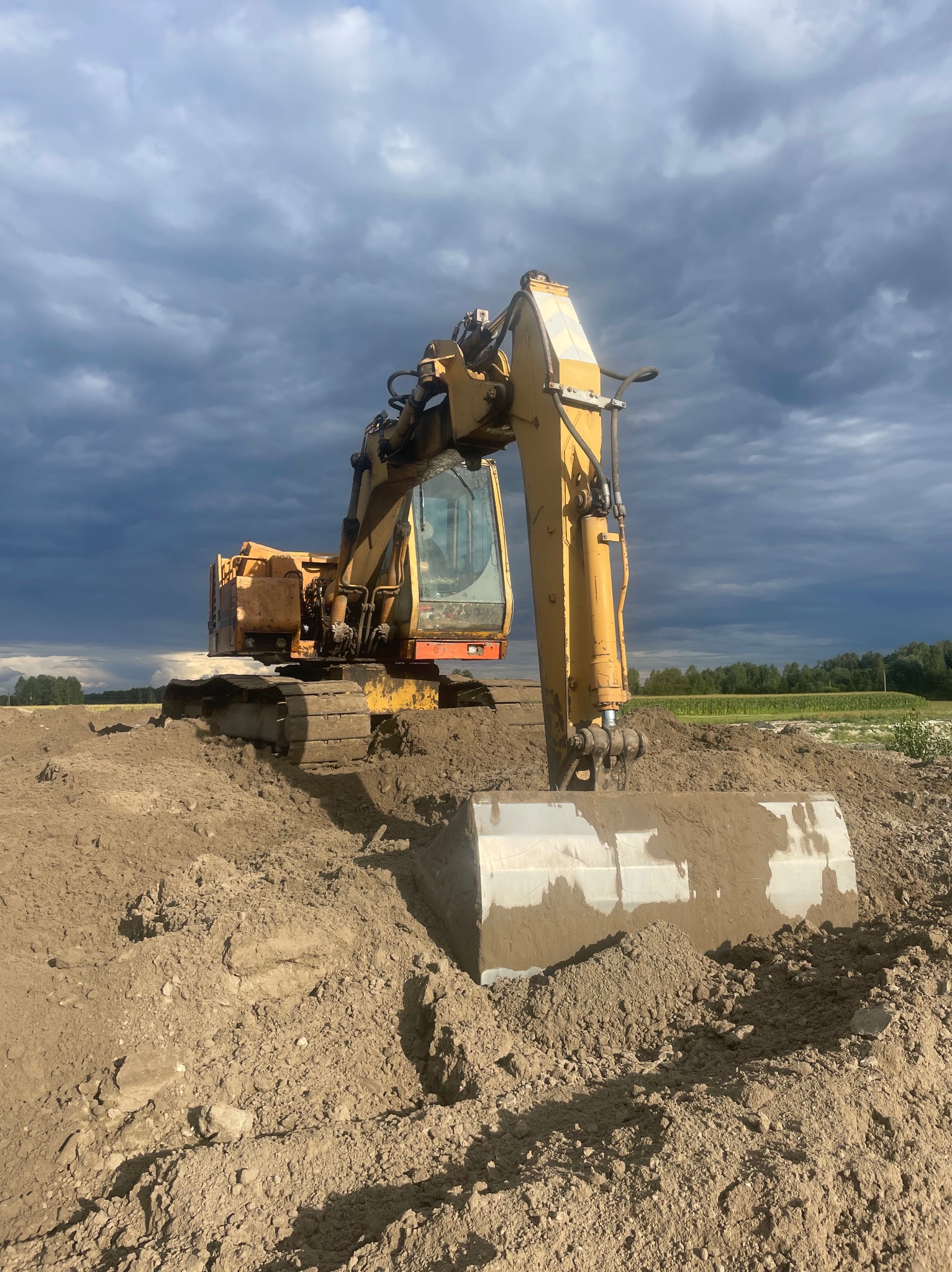 Żółta koparka na placu budowy, z widocznym nabranym gruntem w łyżce, na tle pochmurnego nieba i zieleni w oddali. Maszyna gotowa do pracy.