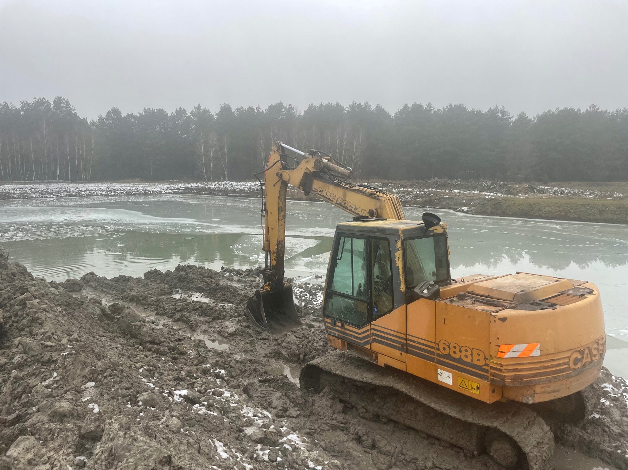 Żółta koparka CASE 688B na błotnistym terenie, obok częściowo zamarzniętego zbiornika wodnego. W tle widoczny las w zimowej scenerii.