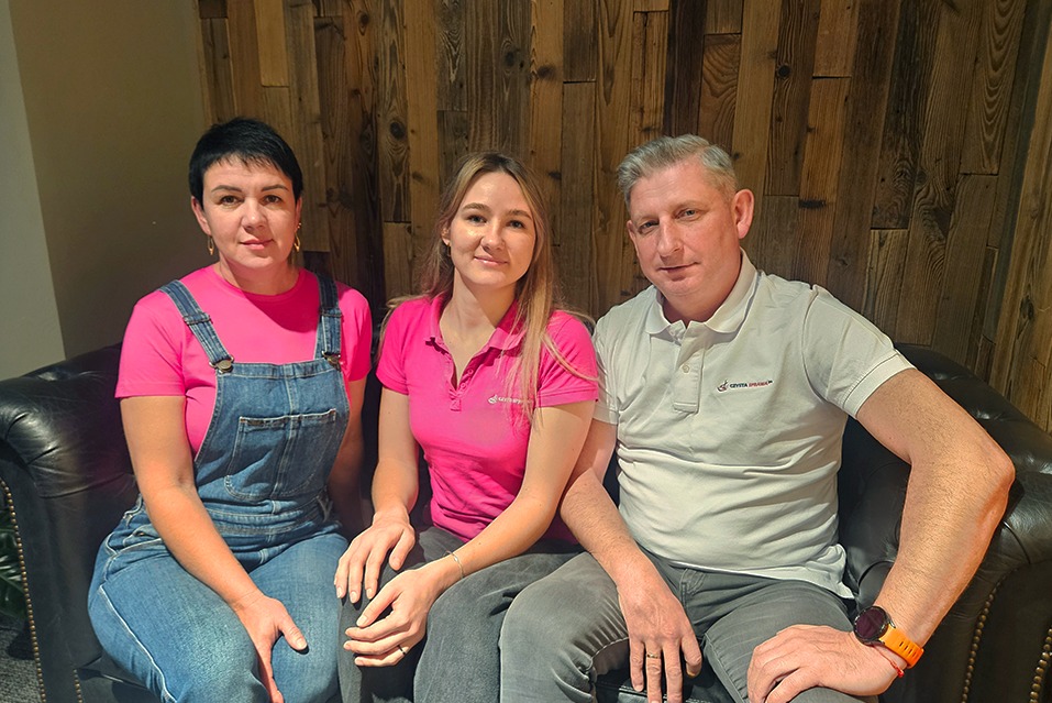 Trzy osoby w różowych koszulkach i dżinsach siedzą na skórzanej kanapie na tle drewnianej ściany. Dwie osoby mają logo firmy sprzątającej na koszulkach.